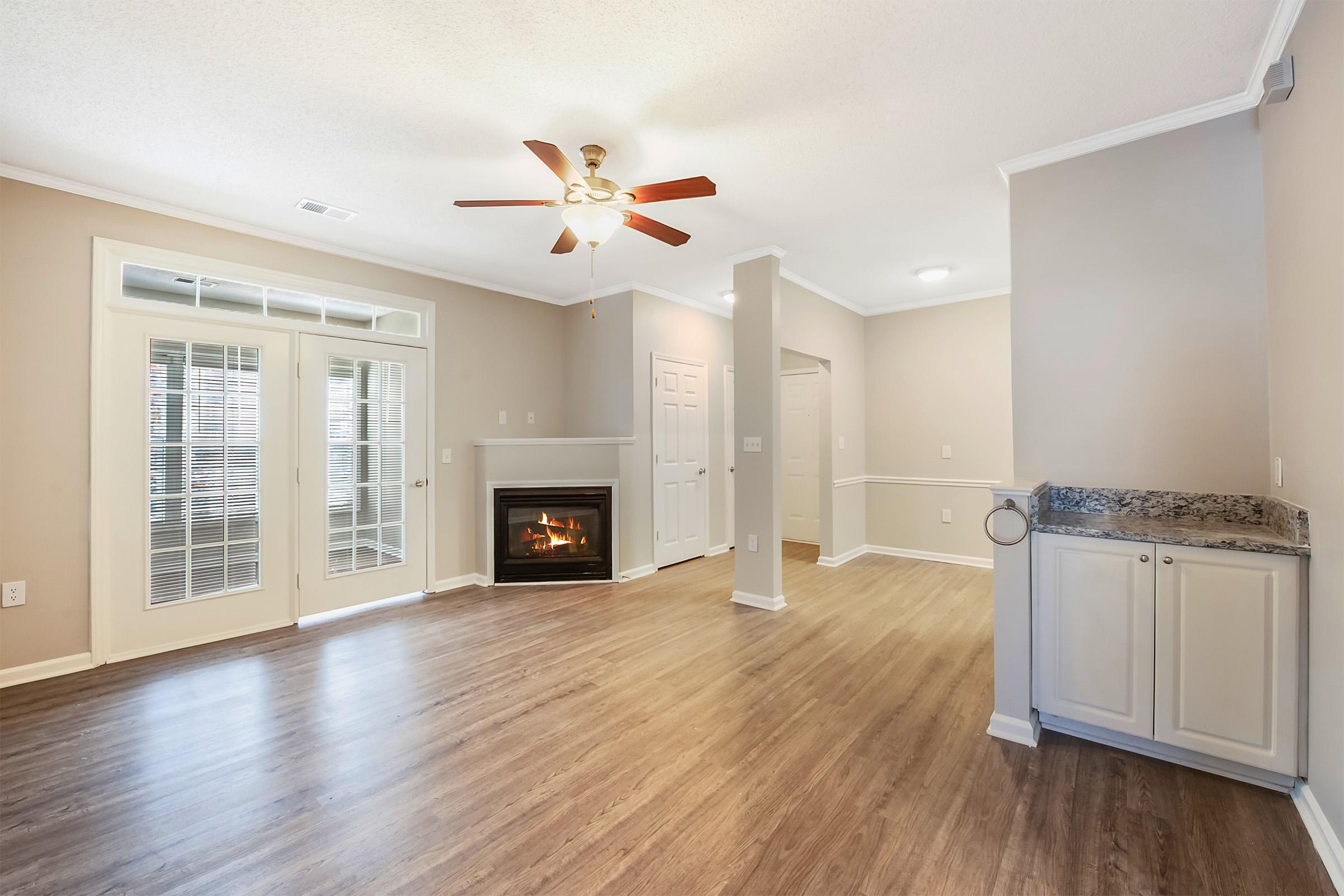 A bright, spacious living room featuring a hardwood floor, a ceiling fan, and a cozy fireplace. There are closed double doors leading to an outdoor space and a small wet bar area. Neutral-colored walls enhance the open, airy feel of the room, creating a welcoming atmosphere.