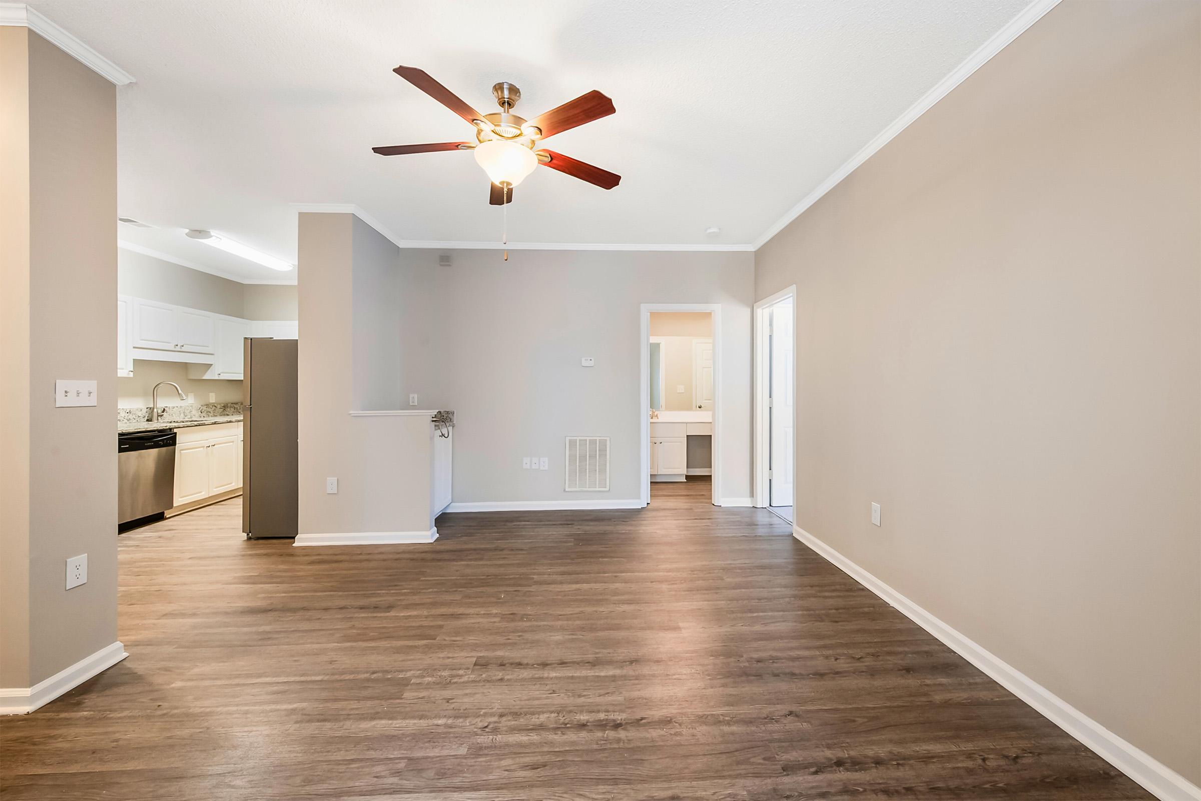 Spacious living area featuring a ceiling fan, light-colored walls, and wood-style flooring. A doorway leads to a bathroom visible in the background, and the kitchen is partially shown with modern appliances. The open layout creates a bright and inviting atmosphere.