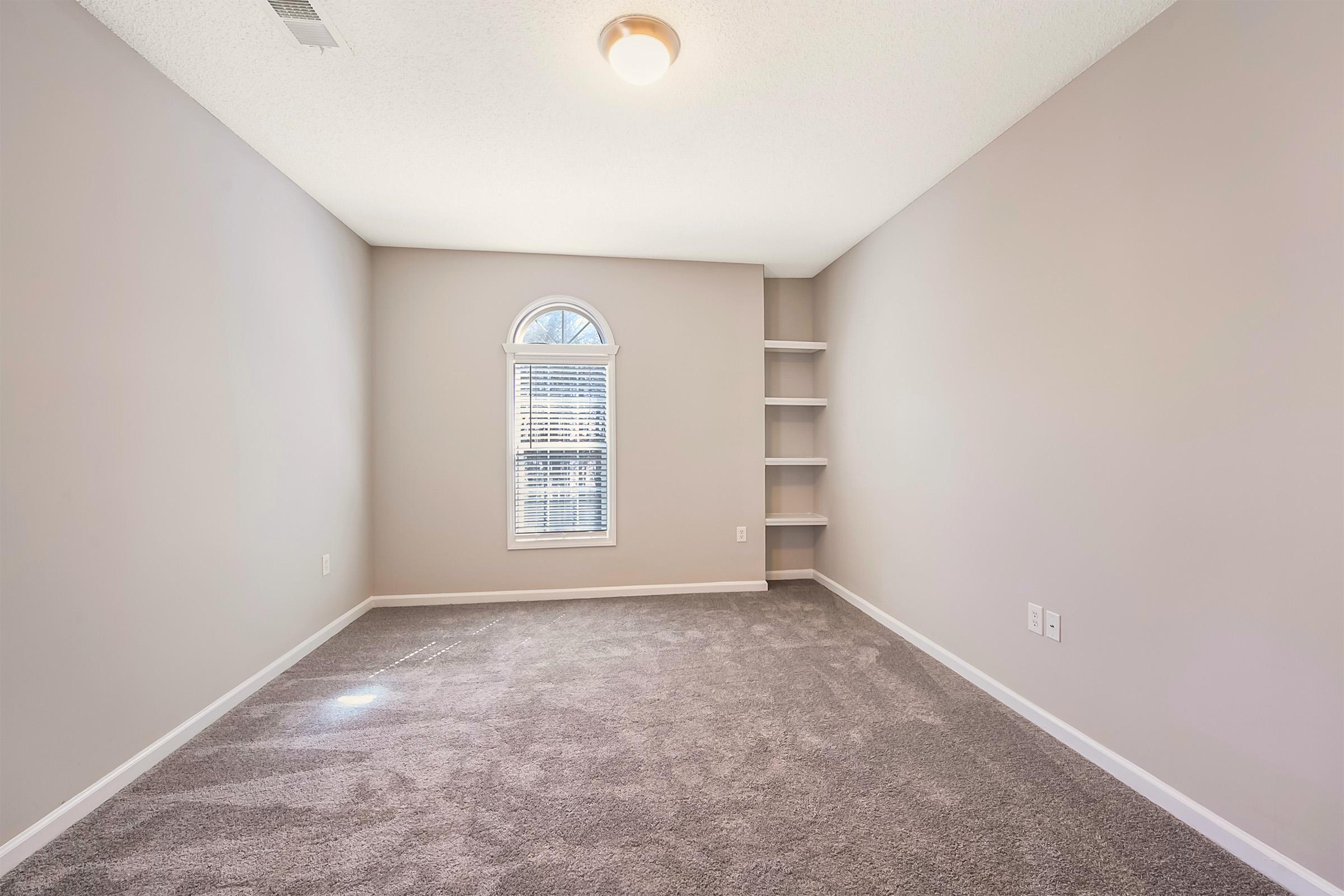 A spacious, empty room featuring light gray walls, plush carpet, and a single arched window with white blinds. A small shelf unit is visible on the right wall, and the ceiling has a standard light fixture. The overall ambiance is bright and neutral, suitable for various decorating styles.