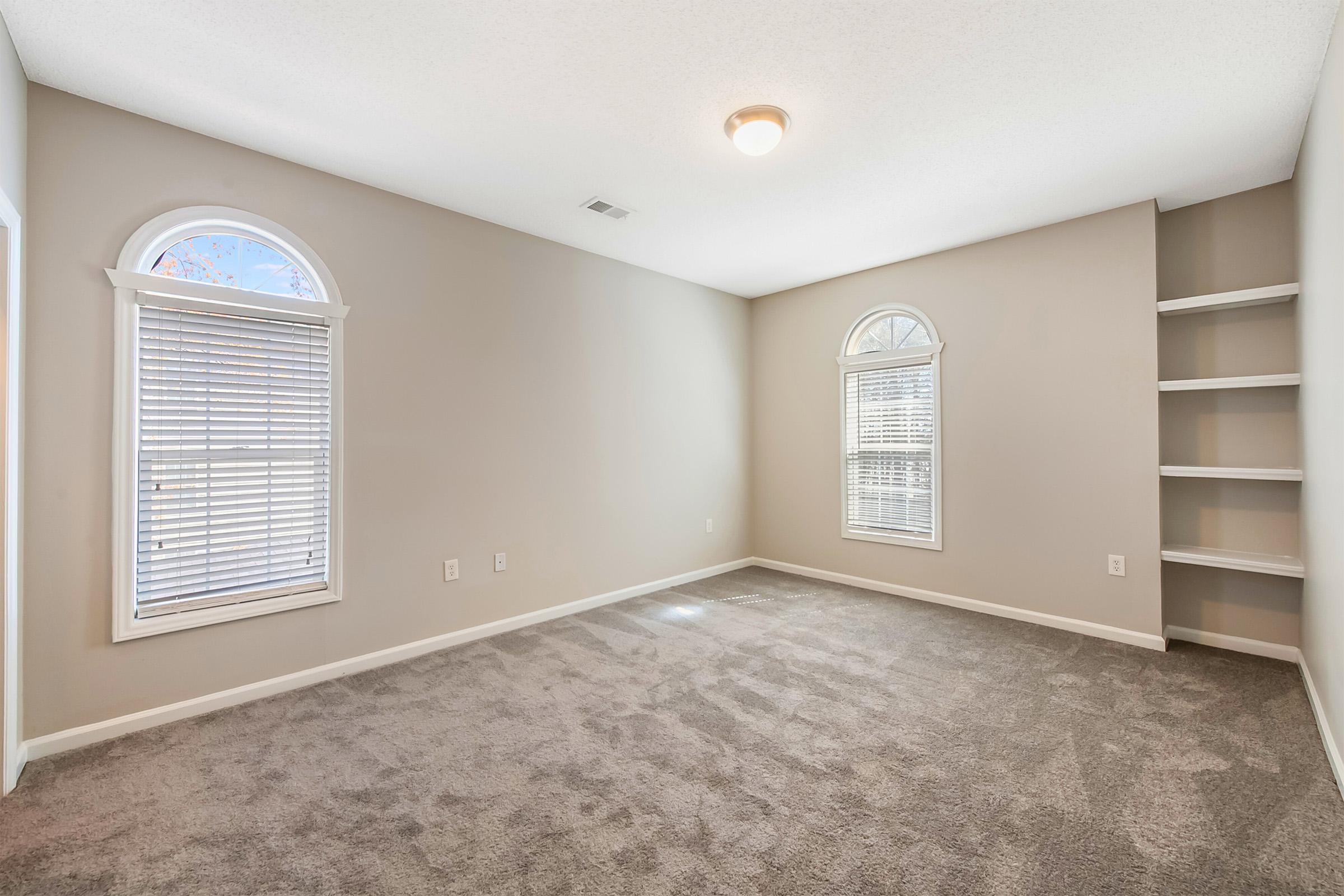 A well-lit, empty bedroom featuring two arched windows with blinds, light gray walls, and a carpeted floor. There is a small niche with shelves on the right side. The ceiling light provides additional illumination, creating a spacious and airy atmosphere.