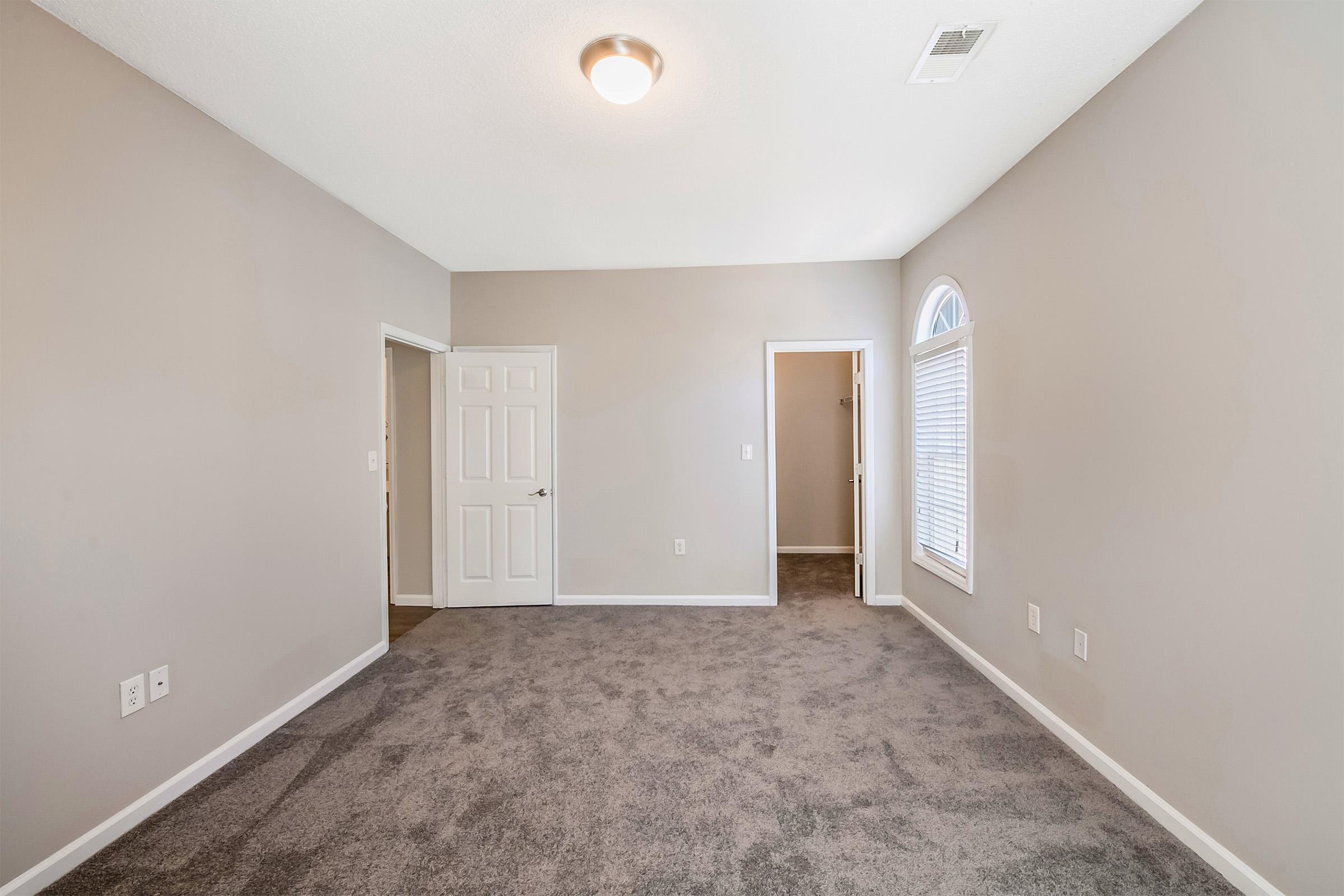 A spacious, well-lit room with gray walls and carpeted flooring. There's a door on the left leading to another room and a closet, while a window on the right allows natural light to enter. The ceiling features a simple light fixture, enhancing the airy feel of the space.