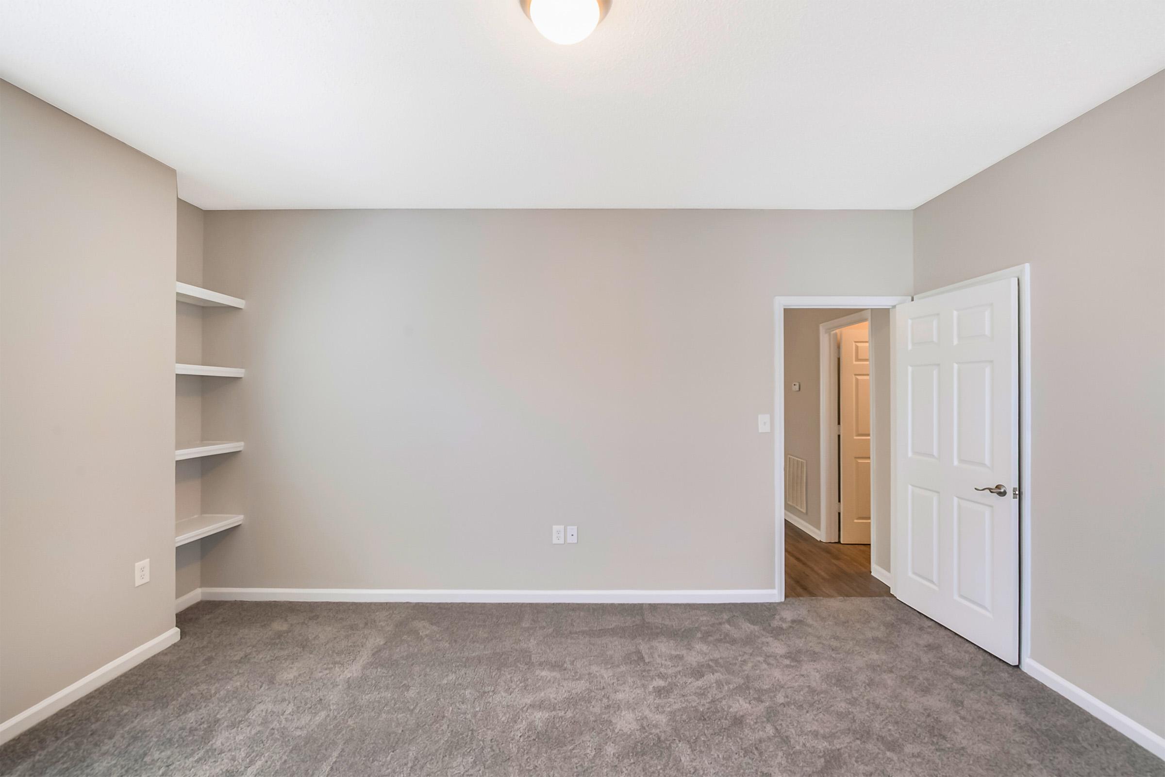 A clean, empty room with light gray walls and carpeted flooring. One wall features open shelves, while a door on the right leads to another room. A ceiling light provides illumination, creating a bright and airy atmosphere. Ideal for various uses like an office or guest room.