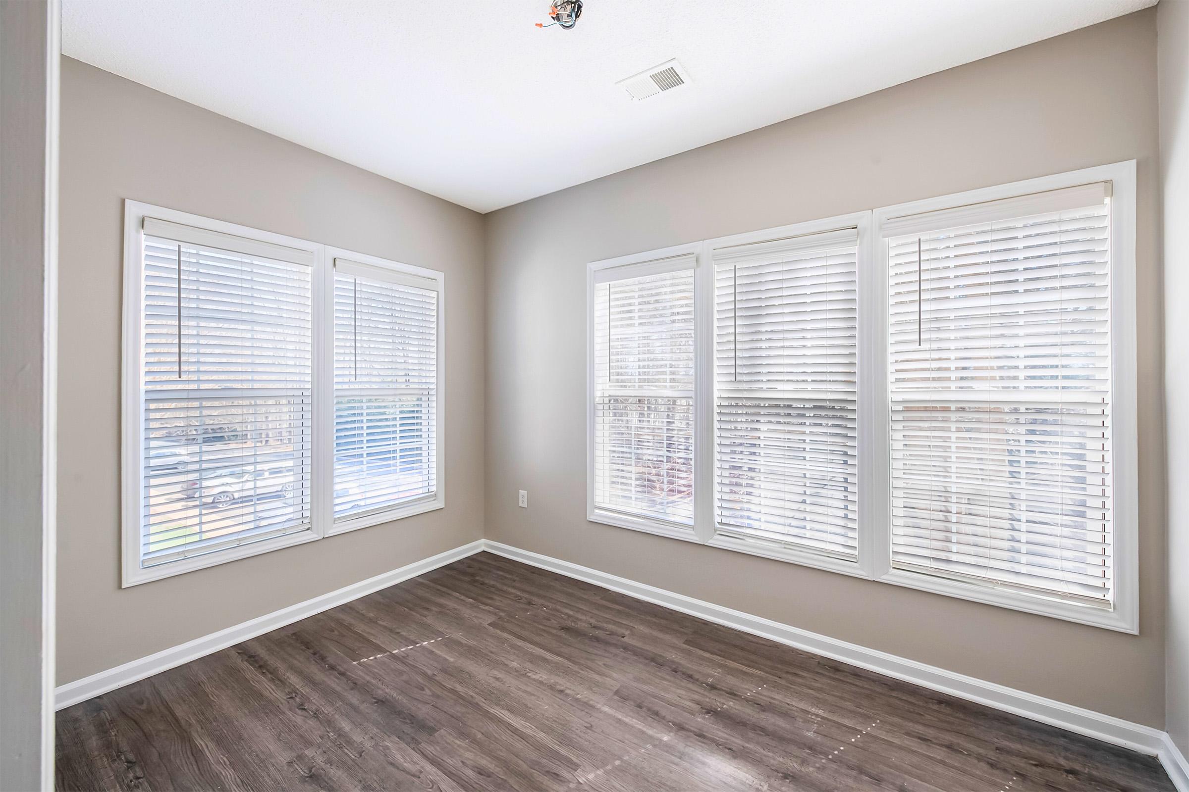 Empty room with light-colored walls and large windows featuring white blinds. The flooring is dark wood, and there is a small ceiling fixture in the center. The sunlight is streaming through the windows, illuminating the space.
