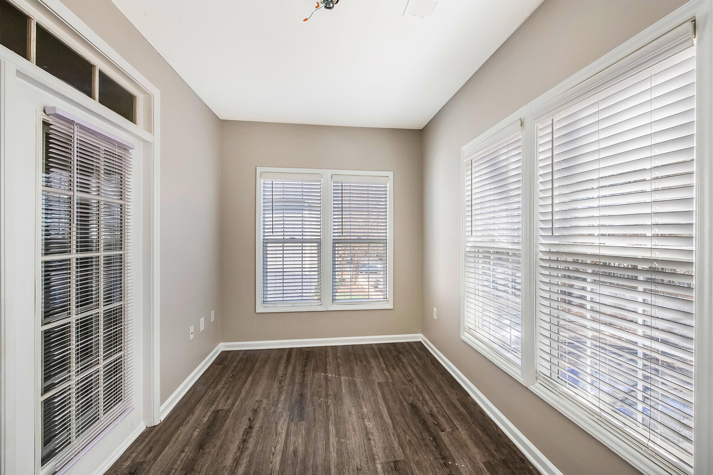 A small room with hardwood flooring and light beige walls, featuring large windows with blinds allowing natural light. The room has a ceiling fan and is empty, creating a spacious atmosphere.