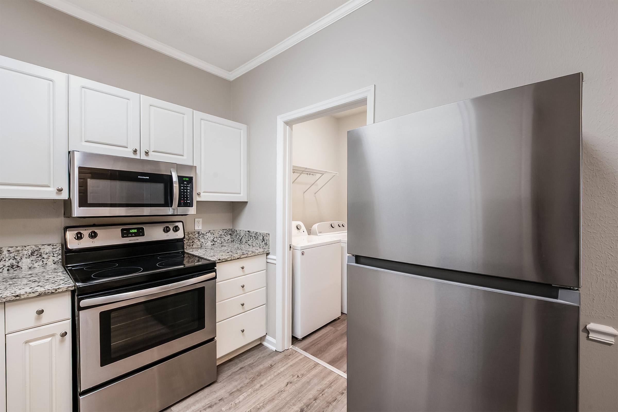 A modern kitchen featuring stainless steel appliances, including a microwave, oven, and refrigerator. White cabinets with granite countertops and light-colored walls create a clean and inviting atmosphere. An adjacent doorway leads to a laundry area with a washer and dryer.