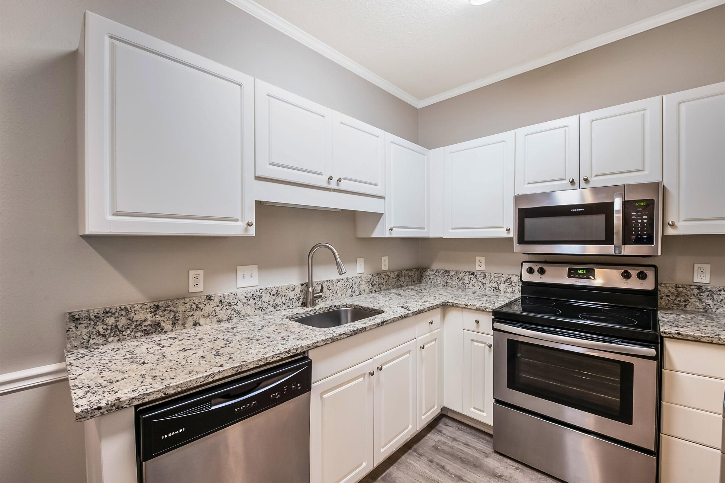 Modern kitchen featuring white cabinetry, granite countertops, stainless steel appliances, including a microwave, oven, and dishwasher. The walls are painted a soft gray, and the space is well-lit by overhead lighting. The layout is functional with a focus on clean lines and a contemporary aesthetic.
