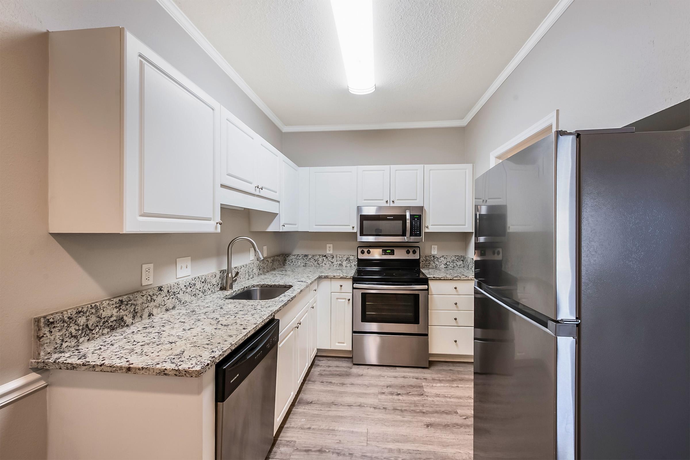 Modern kitchen featuring white cabinetry, granite countertops, and stainless steel appliances, including a microwave, oven, and dishwasher. The space is well-lit with a ceiling fixture and has a neutral color scheme, complemented by wood-like flooring.