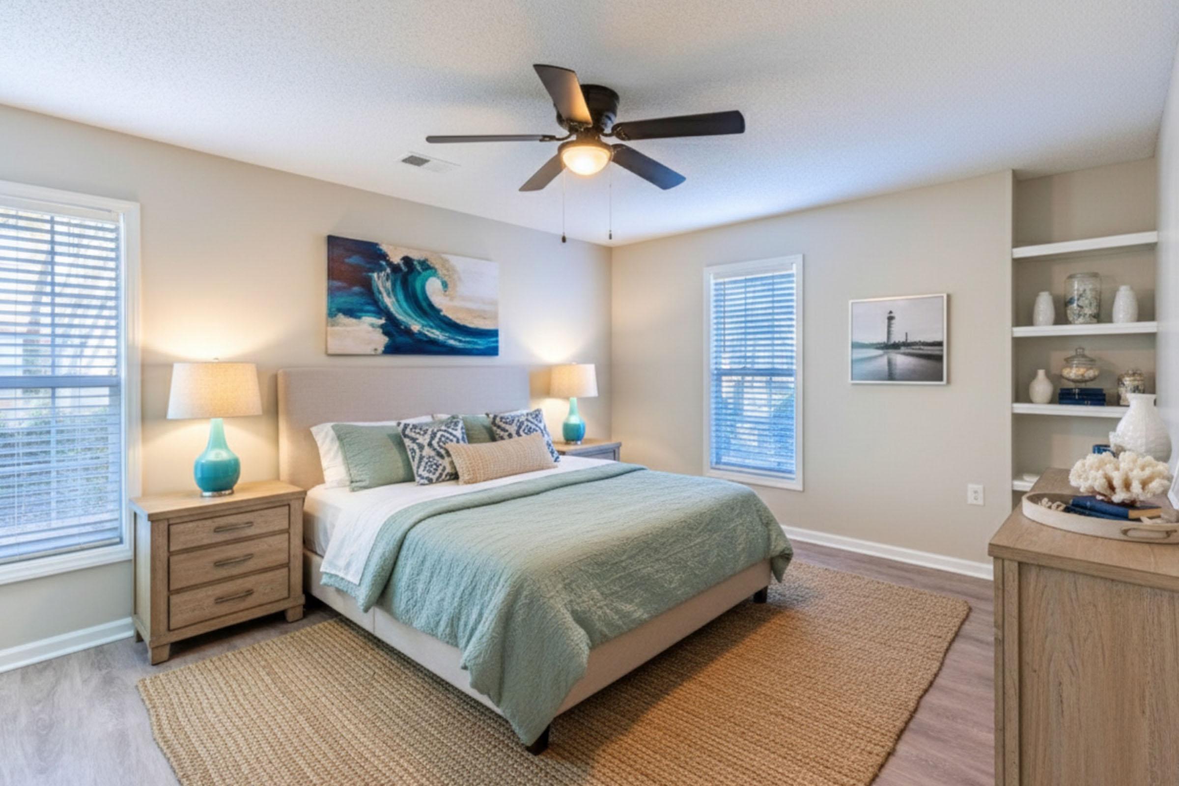 A neatly arranged bedroom featuring a large bed with light blue bedding, two bedside tables with lamps, and a decorative wall painting. There’s a ceiling fan, windows with blinds letting in natural light, and a cozy rug on the floor. A bookshelf displays decorative items, adding a touch of style to the room.