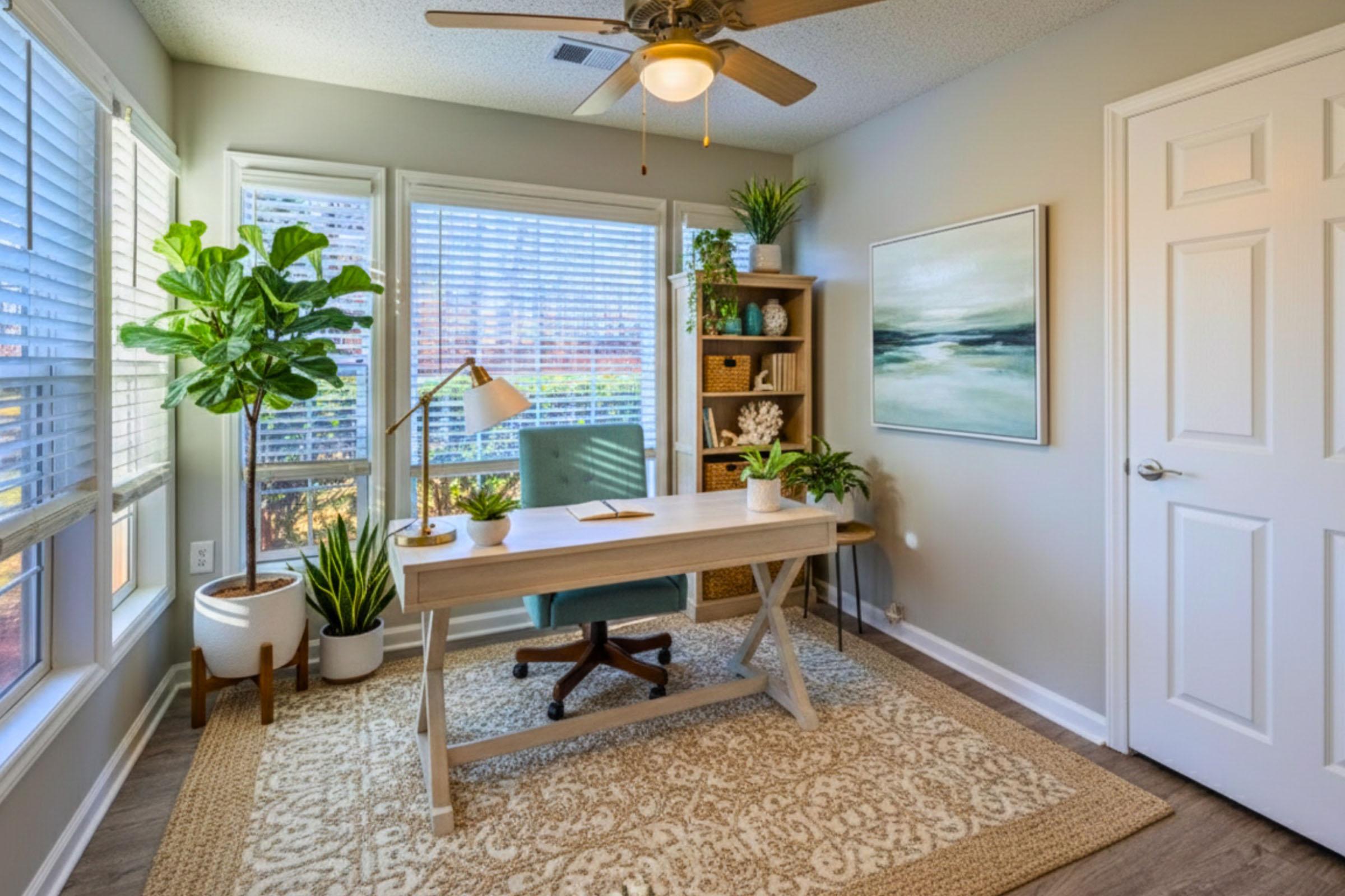 A bright and airy home office featuring a wooden desk with a green chair, a floor lamp, and decorative plants. Large windows allow plenty of natural light, highlighting a cozy rug and a wall-mounted artwork. Shelves with plants and decor add a touch of warmth to the space.
