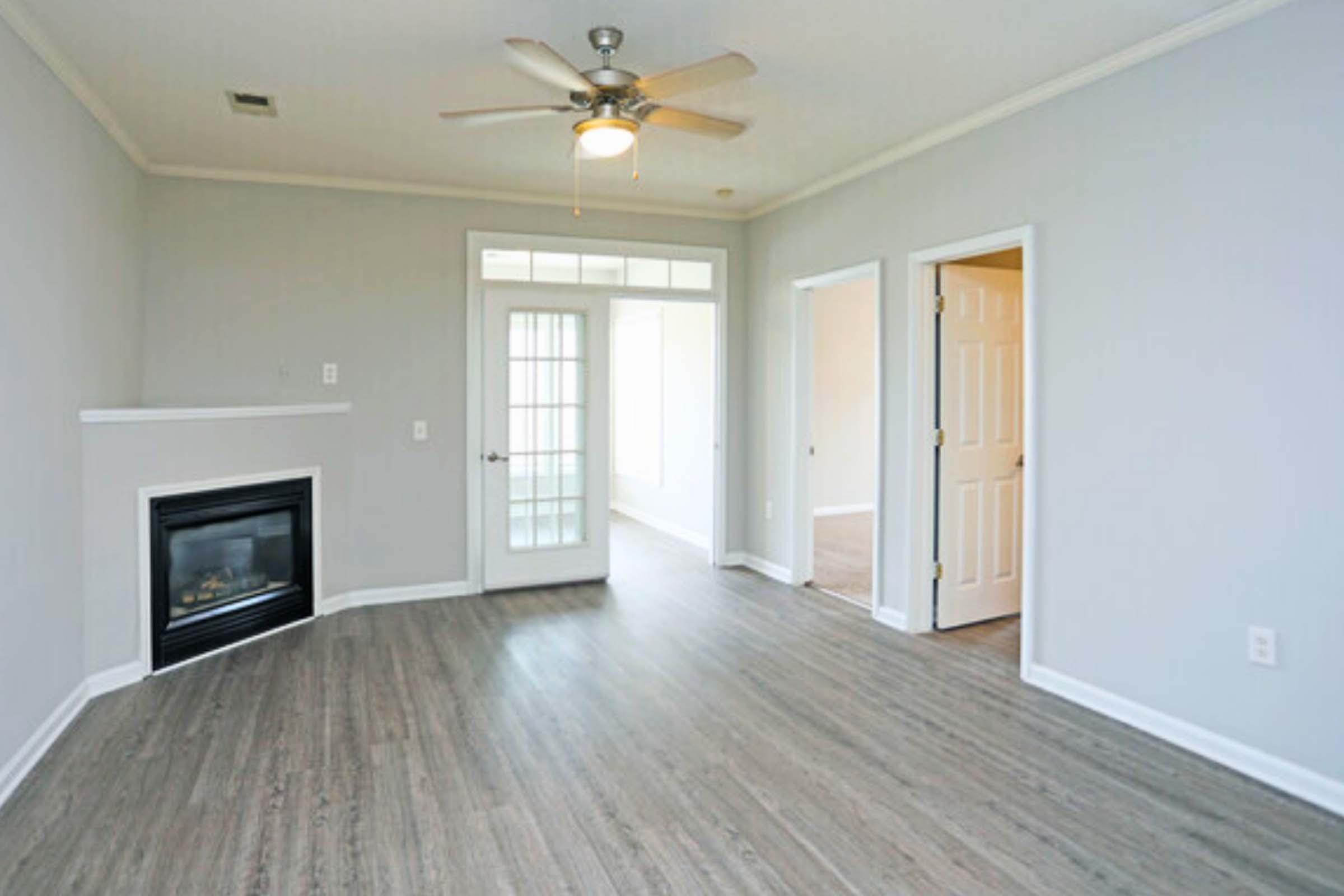 A spacious, empty living room with light gray walls and a ceiling fan. It features a fireplace on the left side and has double glass doors leading to a sunroom. The flooring is light wood, and there are two doorways leading to other rooms. Natural light floods in through the doors.