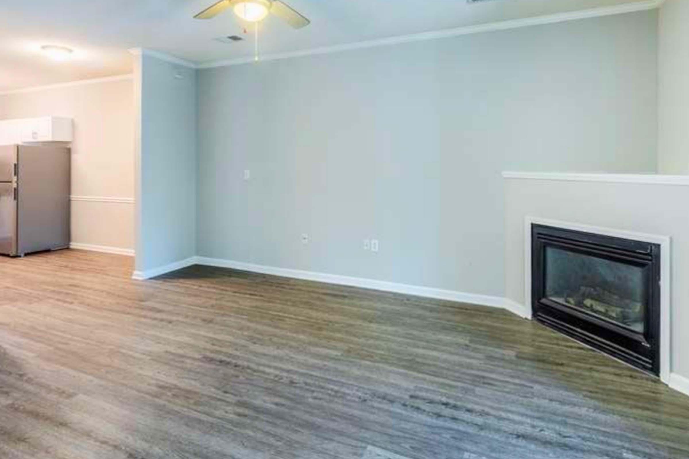 Empty living room featuring wood-like flooring, a modern fireplace on one wall, and a corner kitchen with a stainless steel refrigerator. The walls are painted in a light color and there is a ceiling fan overhead, creating a spacious and airy atmosphere.