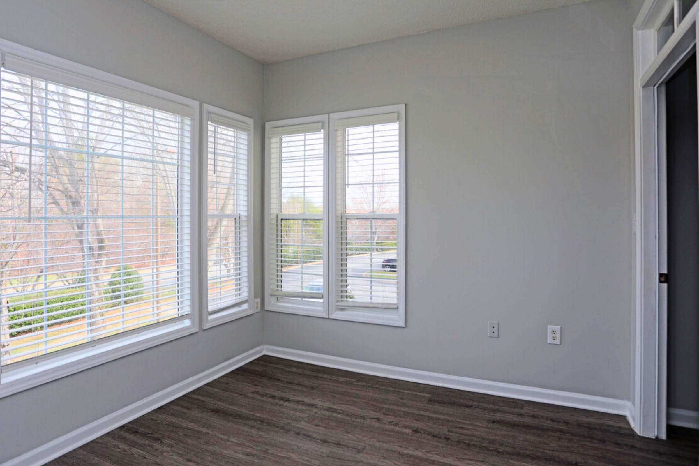 Empty room with gray walls, large windows with white blinds, and wood-style flooring. One corner features a set of triple windows allowing natural light, while another corner shows a doorway. The overall space appears clean and bright, suitable for various uses.