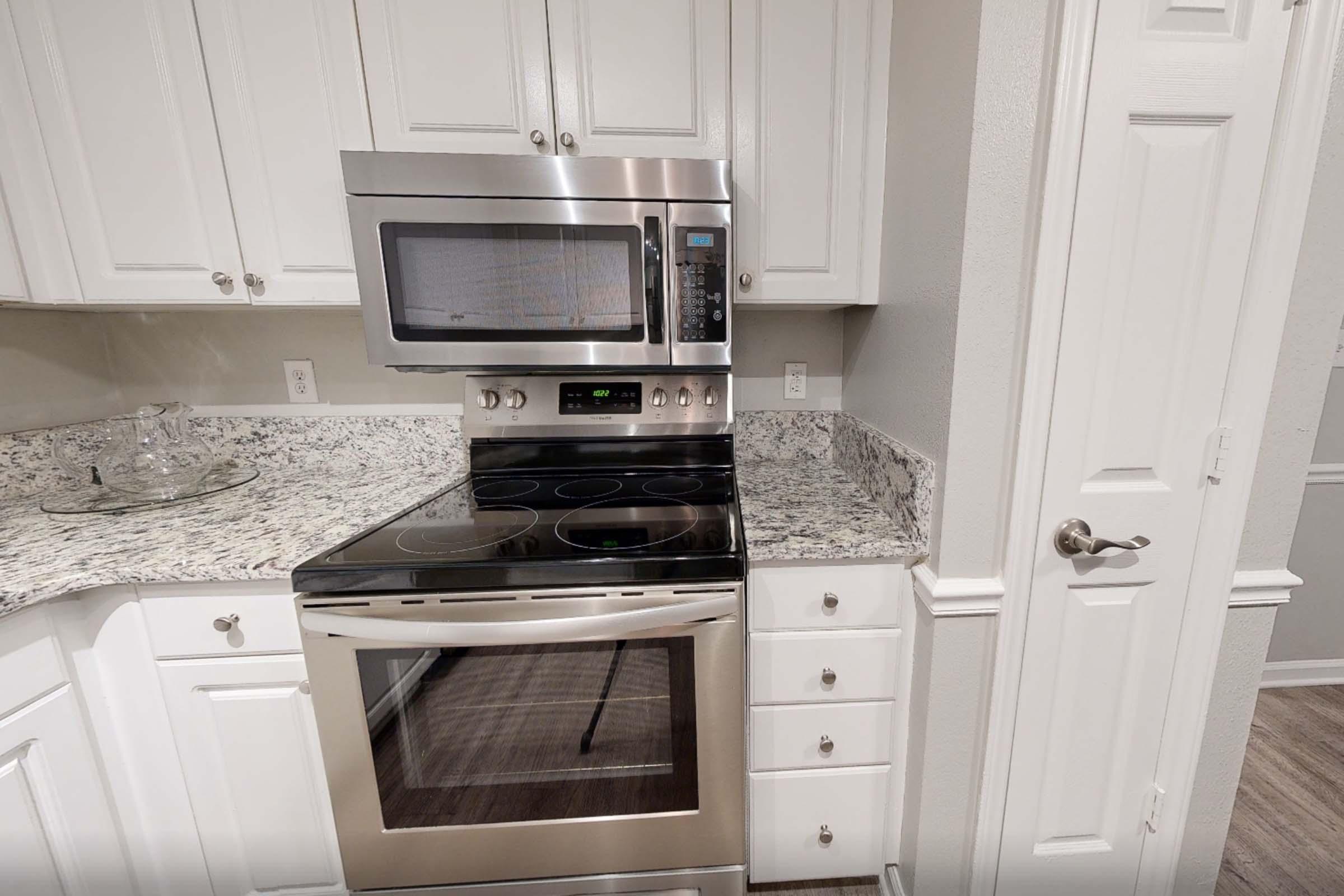 Modern kitchen featuring stainless steel appliances, including an oven and a microwave. The countertops are made of speckled granite, and the cabinets are white. A door on the right leads to another room, while the kitchen area is well-lit and clean.