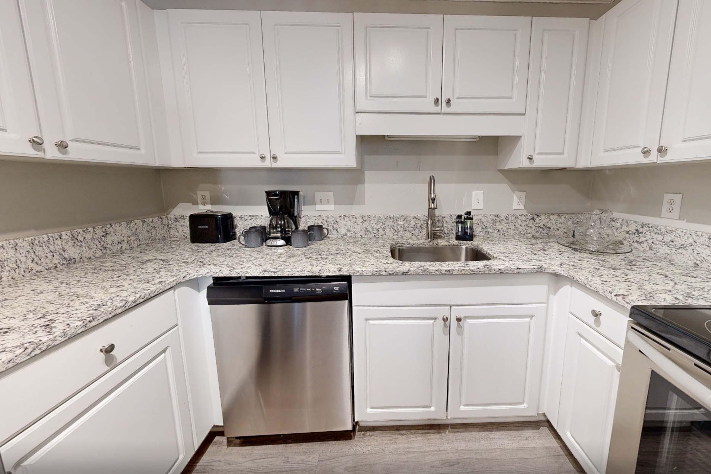 A modern kitchen featuring white cabinets, a granite countertop, a stainless steel sink, a dishwasher, and a coffee maker. The space is well-lit and organized, with various kitchen appliances and utensils neatly arranged on the counter. The flooring is a light wood finish.