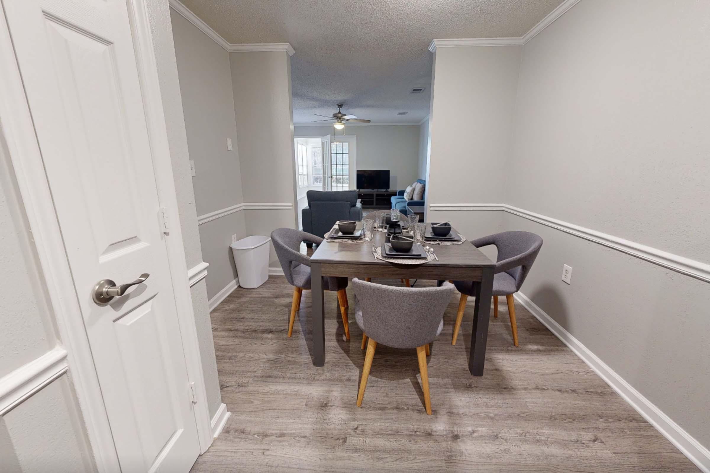 A modern dining area featuring a wooden table set for four with gray upholstered chairs. The space has light gray walls and wood-like flooring, with a trash can nearby. In the background, a living room is visible with a sofa and a ceiling fan. Natural light enters through windows, creating a bright atmosphere.