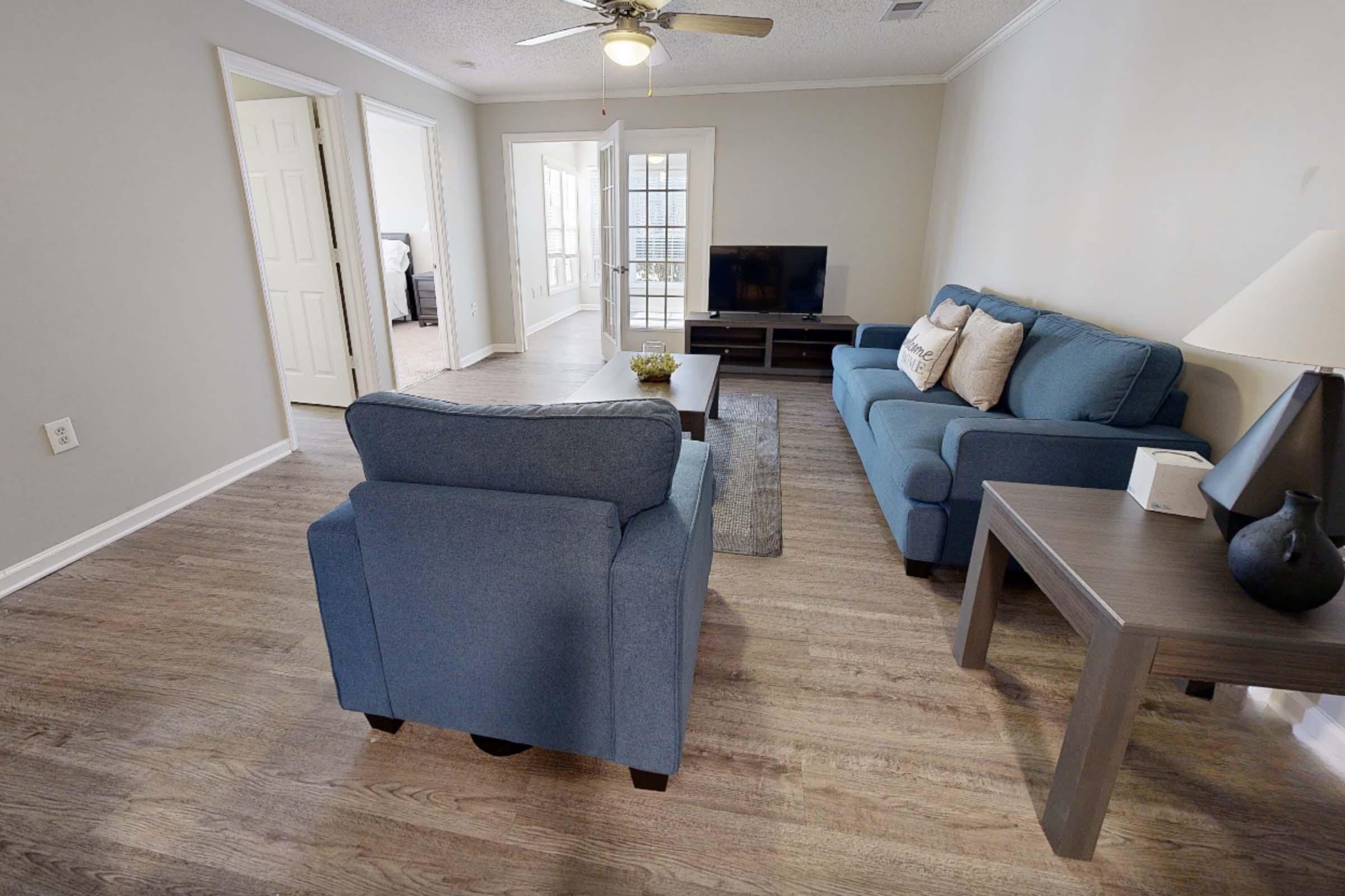 A cozy living room featuring a blue sofa and armchair, a wooden coffee table, and a TV unit. The walls are light-colored, and there's a ceiling fan above. A doorway leads to a well-lit area, and the flooring is wooden, contributing to a warm and inviting atmosphere.