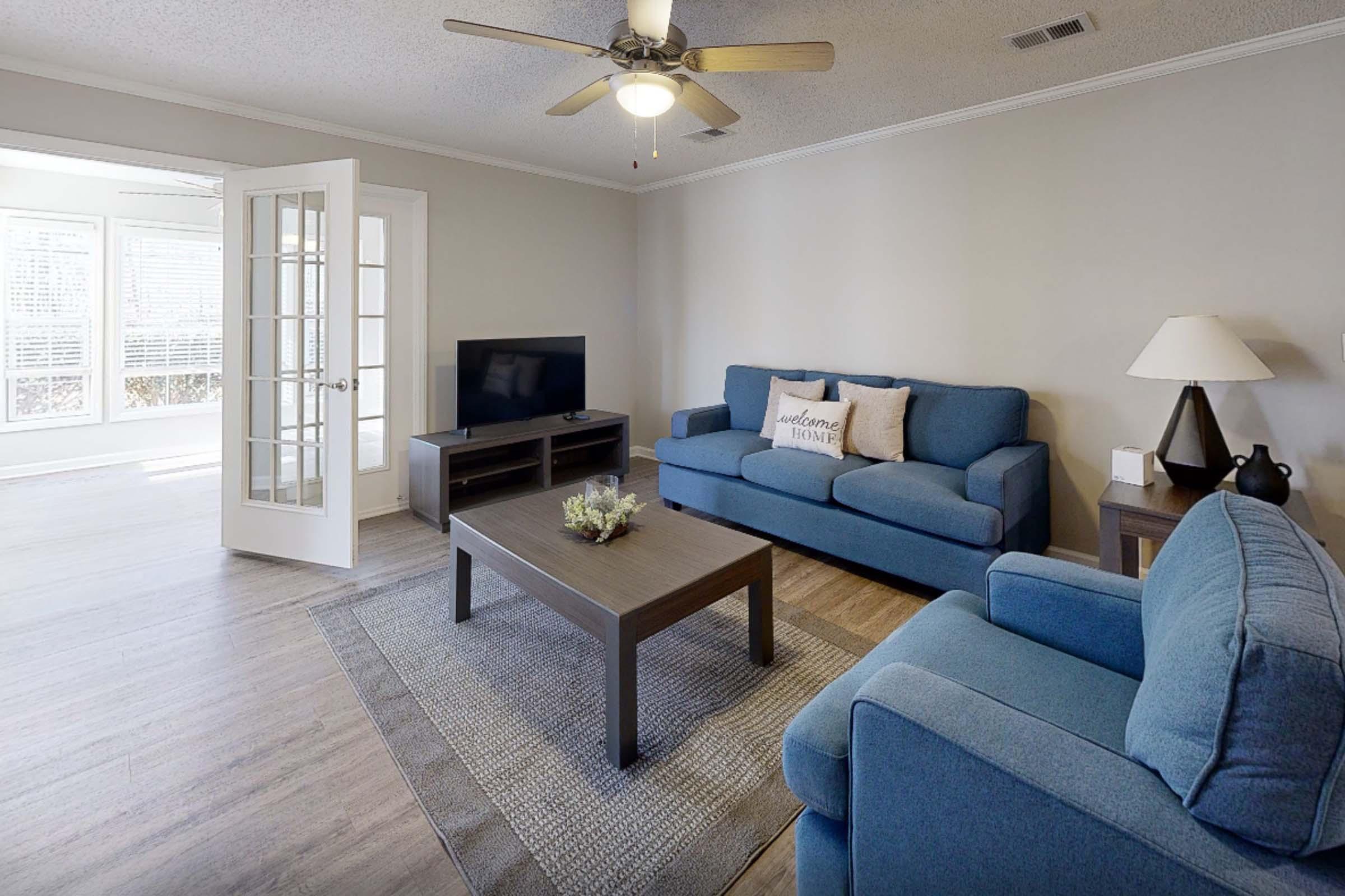 A bright living room featuring a blue sofa and armchair, a wooden coffee table with a small plant, and a television on a stand. There are large windows with natural light and a set of French doors leading to another space, creating a warm and inviting atmosphere.