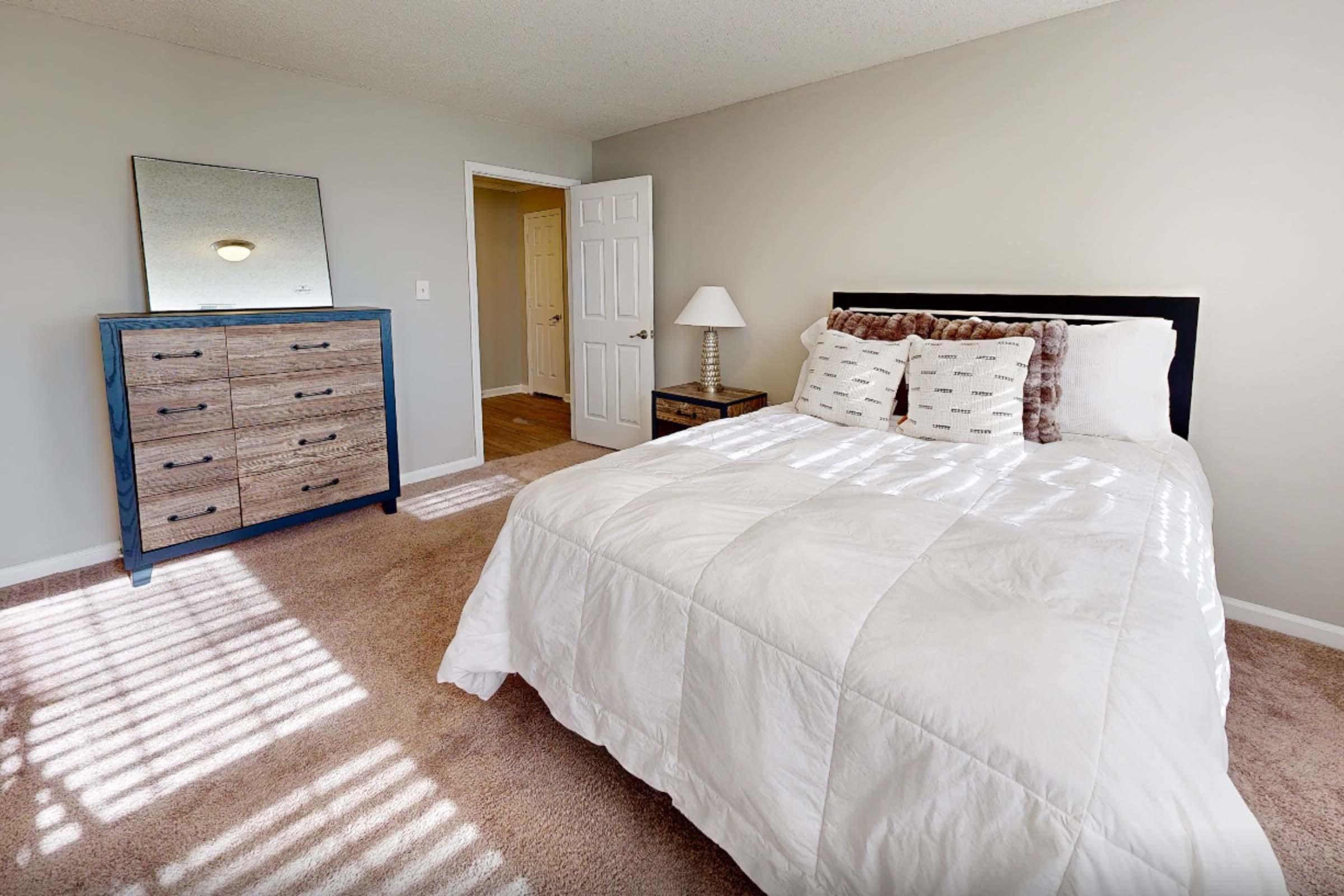 A cozy bedroom featuring a queen-sized bed with white bedding and decorative pillows. There is a wooden dresser with six drawers, a bedside lamp, and a large mirror. Sunlight streams in through a window, casting shadows on the carpeted floor, and an open door leads to a hallway. The walls are painted a light color.