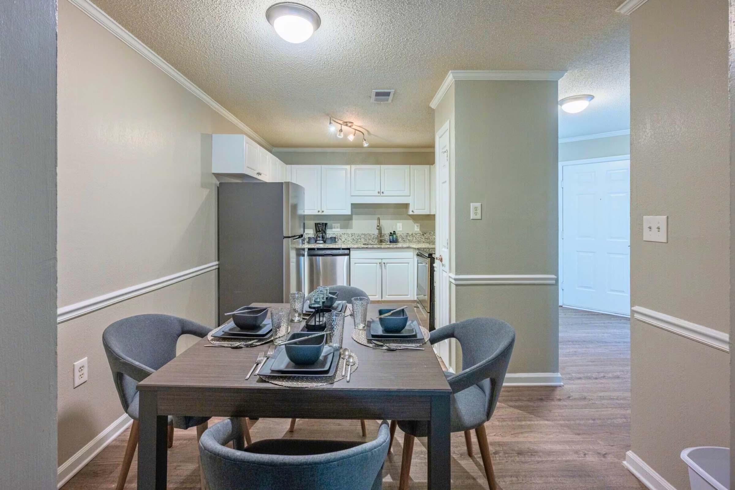 Modern kitchen and dining area featuring a wooden dining table set for four with gray dishes and utensils. The kitchen includes white cabinetry, a stainless steel refrigerator, and light-colored walls. Soft lighting illuminates the space, creating a cozy atmosphere.