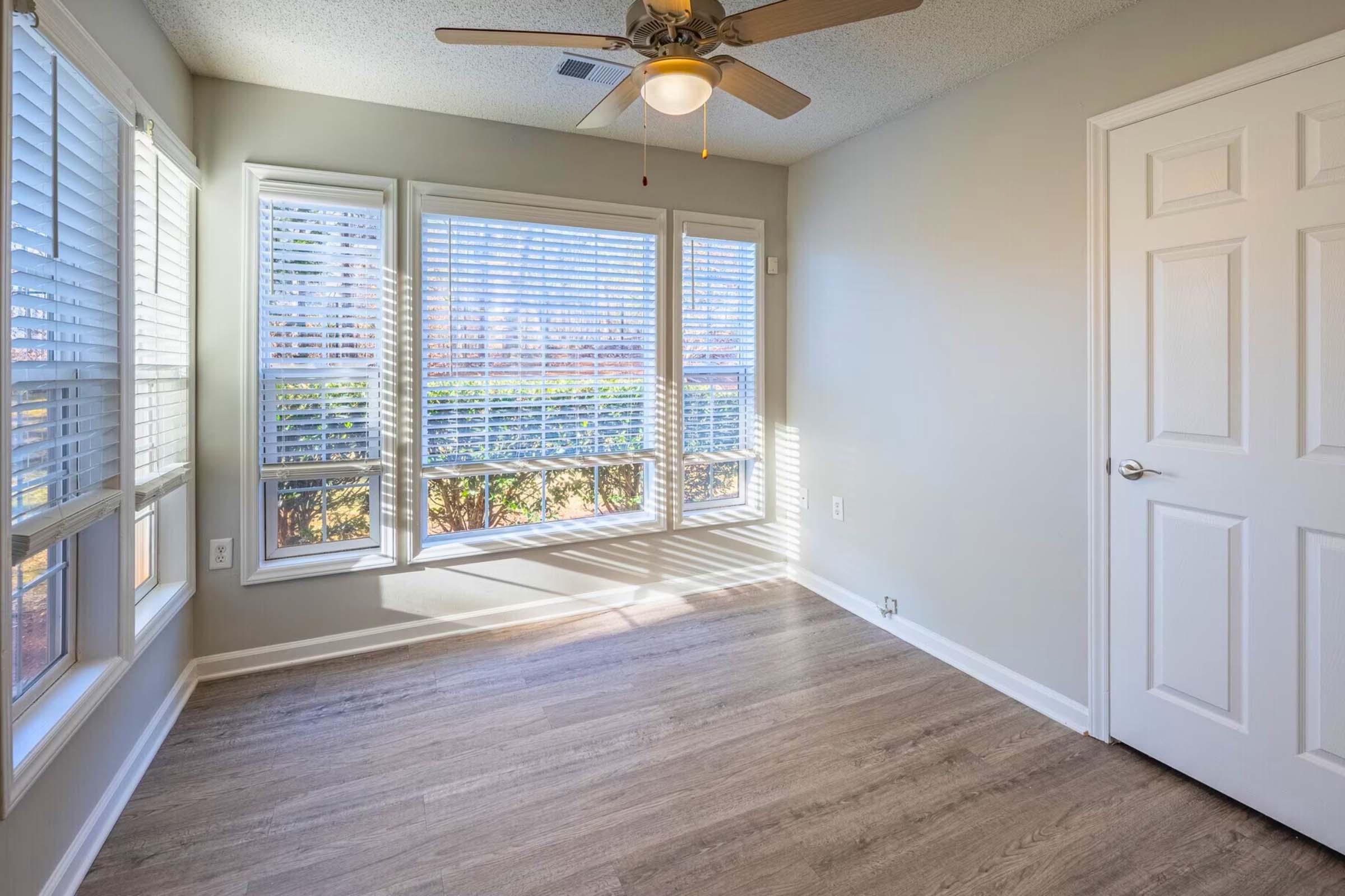 A well-lit, empty room with light-colored walls and a ceiling fan. The room features large windows with white blinds, allowing natural light to filter in, and has laminate flooring. A closed white door is visible on the right side. The overall atmosphere is bright and inviting.