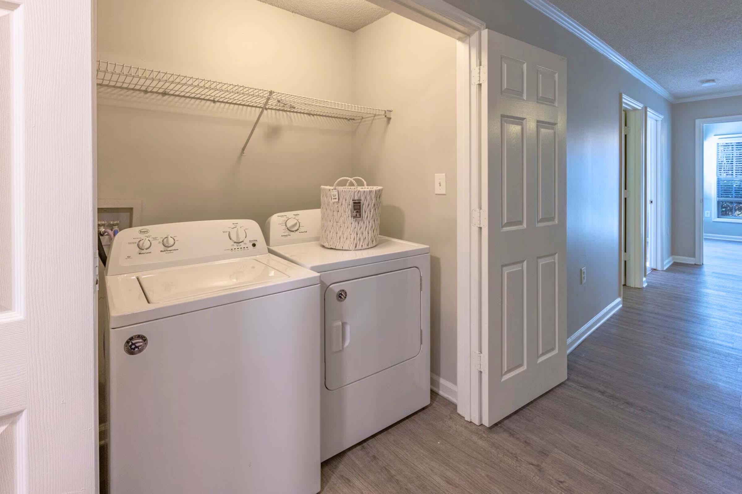 A laundry area featuring a white washing machine and dryer stacked next to each other. Above them is a wire shelf for storage. The room has light gray walls and a doorway leading to a hallway, with a glimpse of a bright living space in the background.