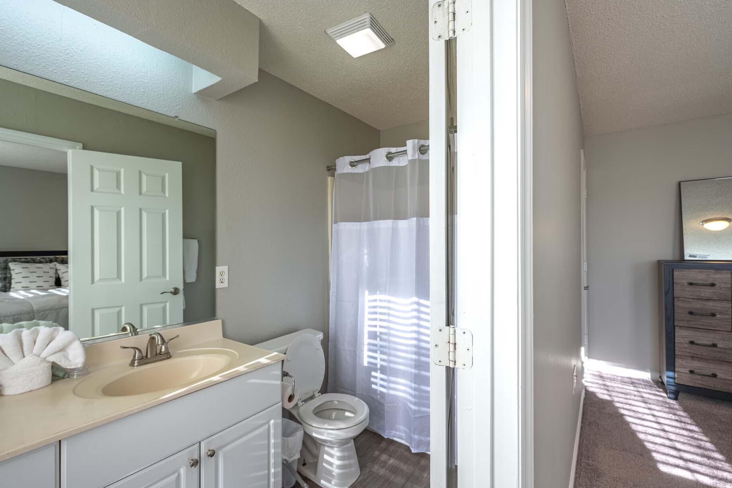 A modern bathroom featuring a sink with light-colored cabinetry, a toilet, and a shower area with a white curtain. The wall color is a soft gray, and there's natural light from a nearby window, casting shadows on the floor. A mirrored cabinet reflects part of the room.