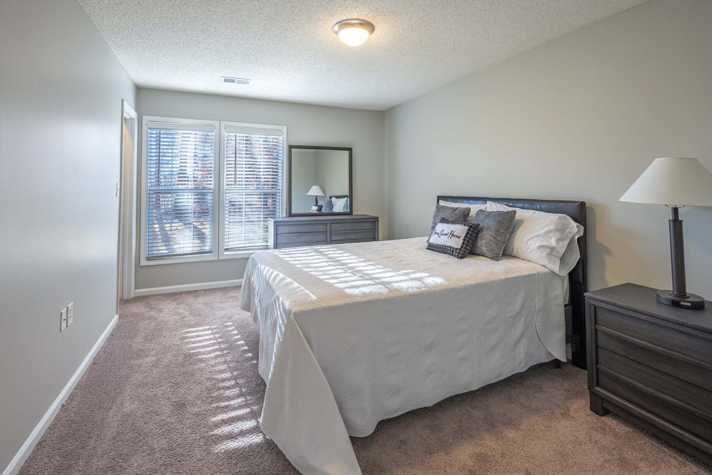 Bright and airy bedroom featuring a queen-sized bed with white bedding, a dark wooden nightstand, and a matching dresser. Natural light streams in through large windows adorned with blinds, casting shadows on the carpeted floor. A lamp sits on the nightstand, and a mirror reflects the space.