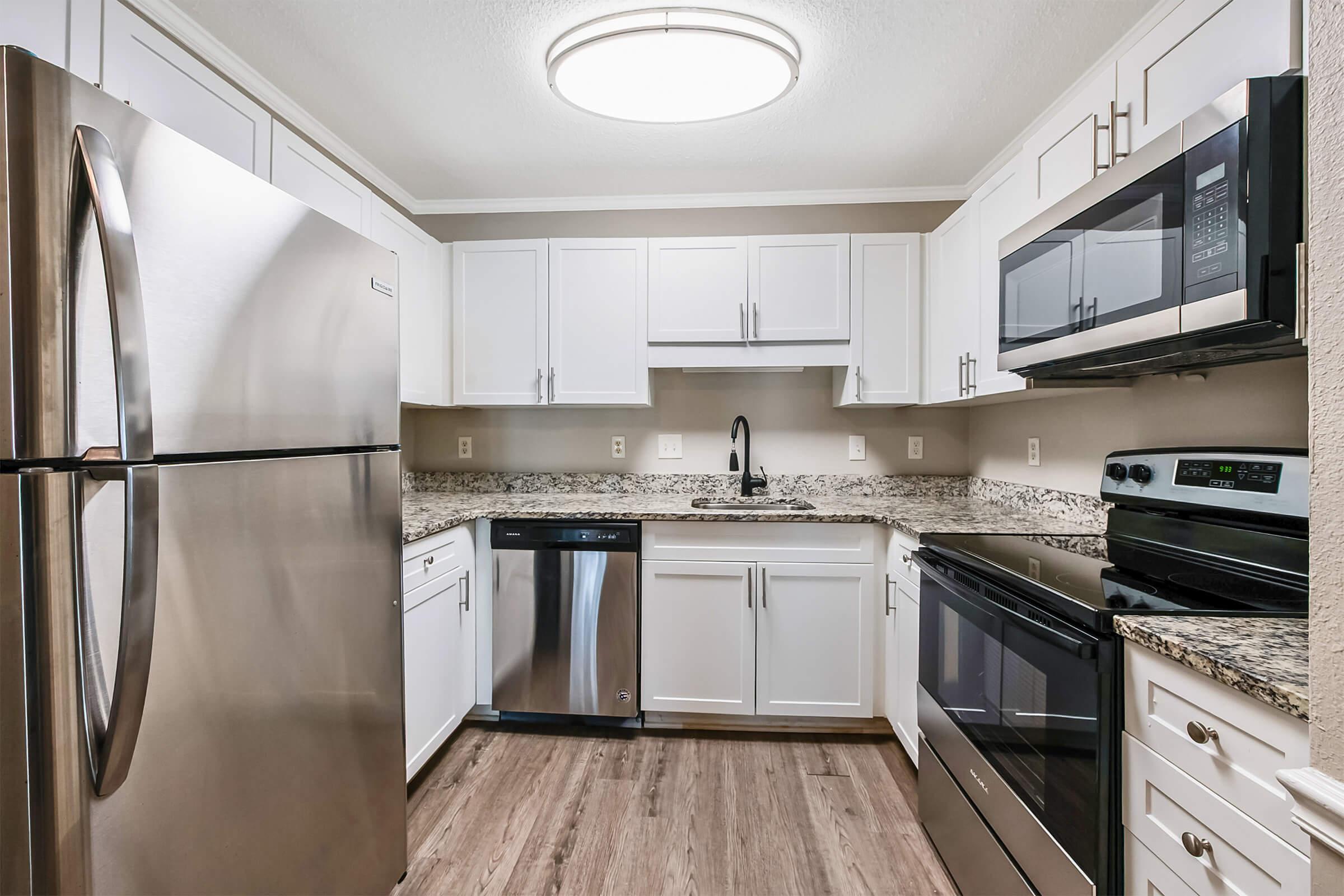 Modern kitchen with white cabinetry, granite countertops, stainless steel appliances including a fridge, dishwasher, microwave, and oven. The space features a ceiling light, neutral wall color, and wood-style flooring, creating a bright and inviting atmosphere.