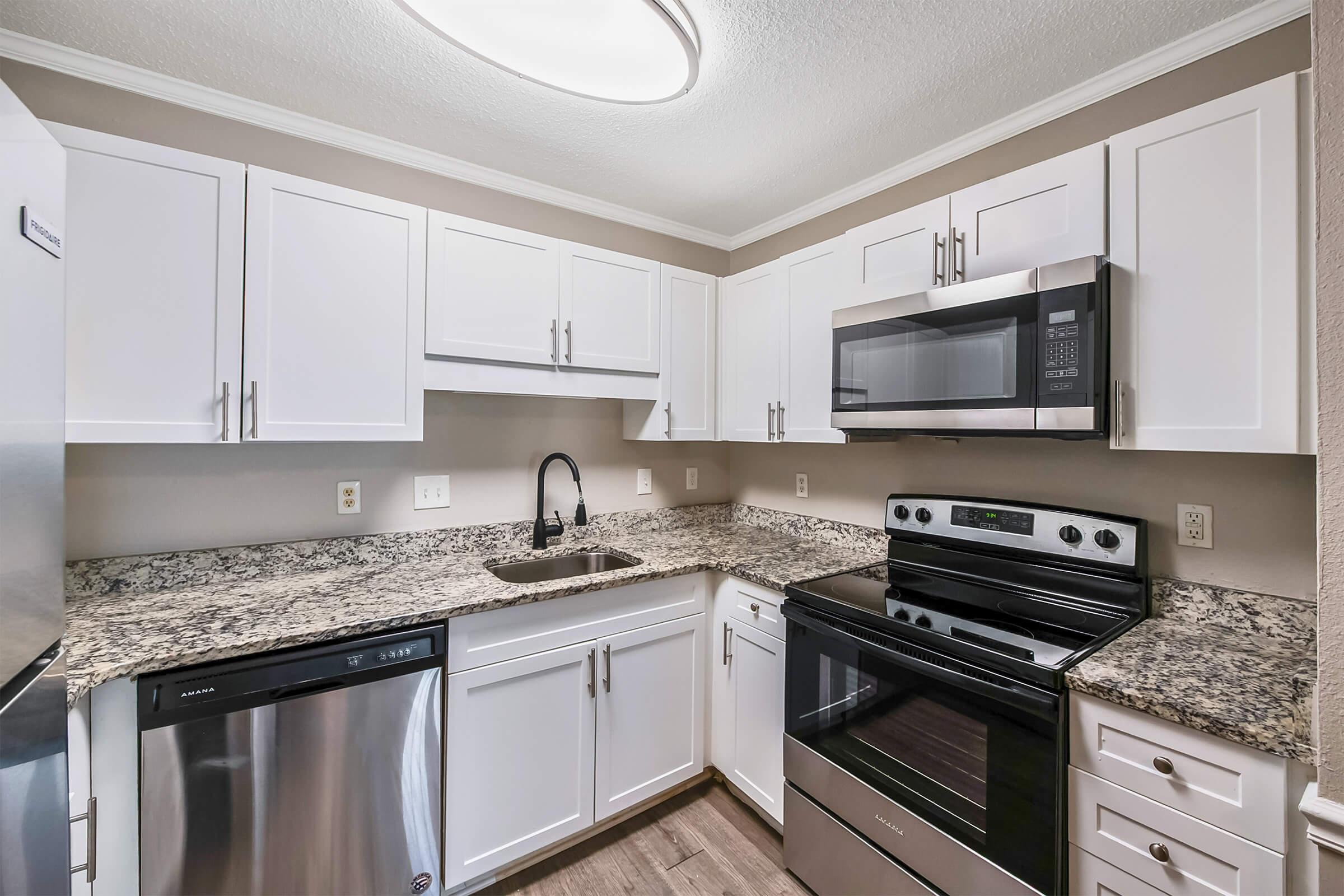Modern kitchen featuring white cabinets, stainless steel appliances including a microwave, oven, and dishwasher, and a granite countertop. The space is well-lit with a circular ceiling light and has a neutral wall color.