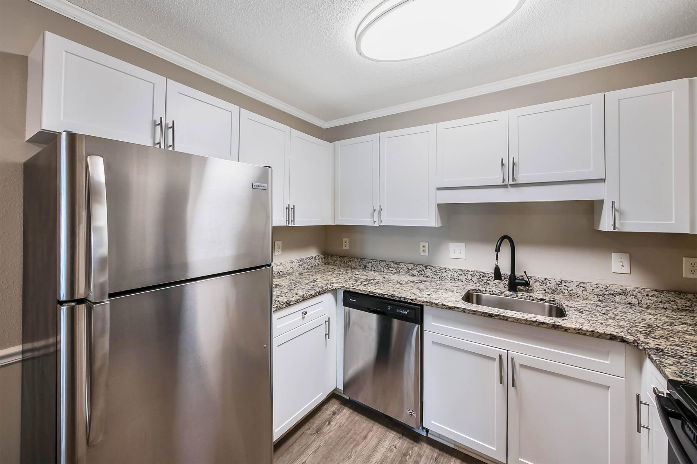 A modern kitchen featuring white cabinets, stainless steel appliances, and a granite countertop. The space includes a refrigerator, dishwasher, and sink with a black faucet, all under a circular ceiling light. The walls are painted in a warm neutral color, enhancing the contemporary design.