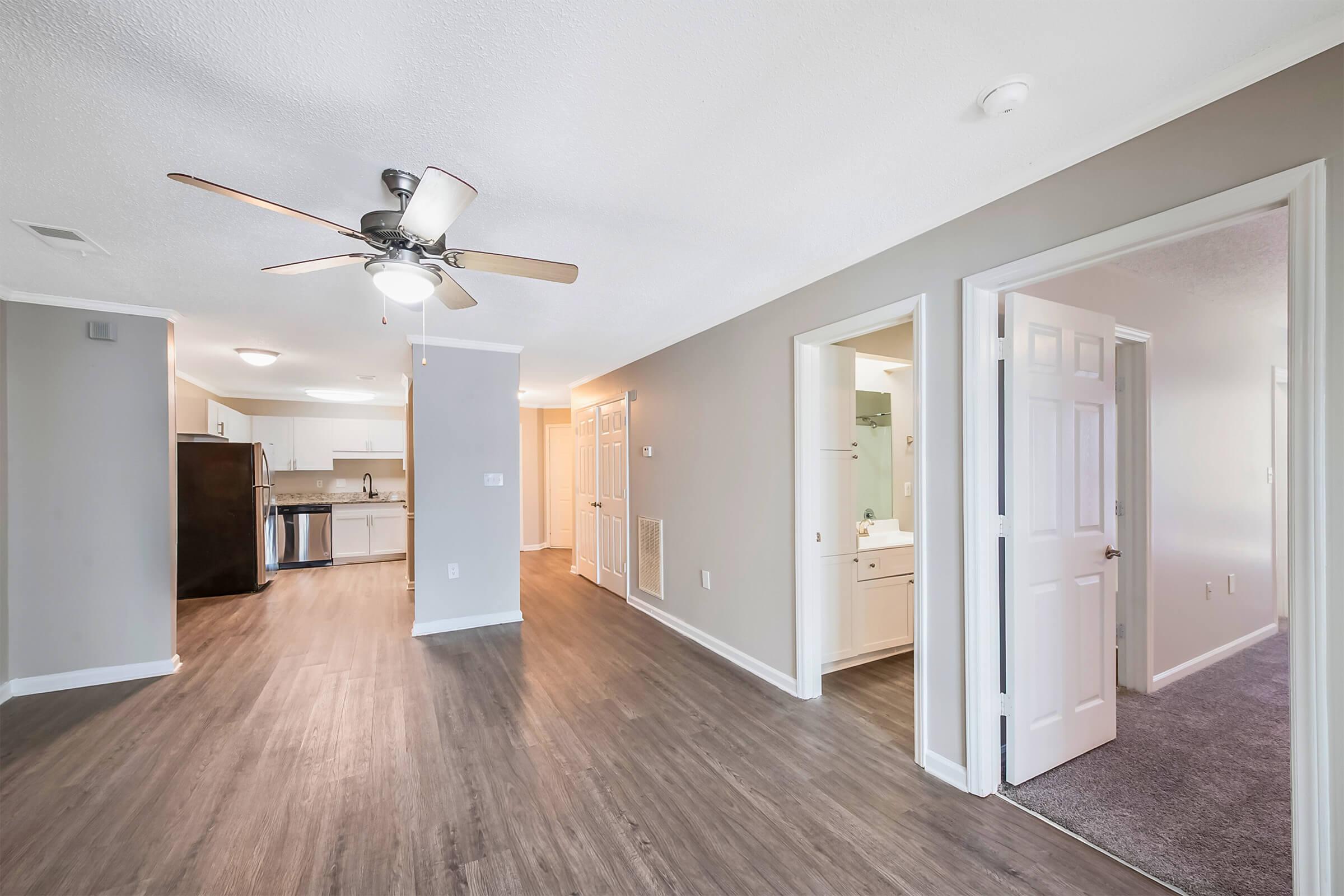 A spacious interior view of an apartment featuring a living area with a ceiling fan, wood-style flooring, and an open layout. Includes doorways leading to other rooms, a kitchenette with appliances visible, and a light-colored wall design creating a modern ambiance.