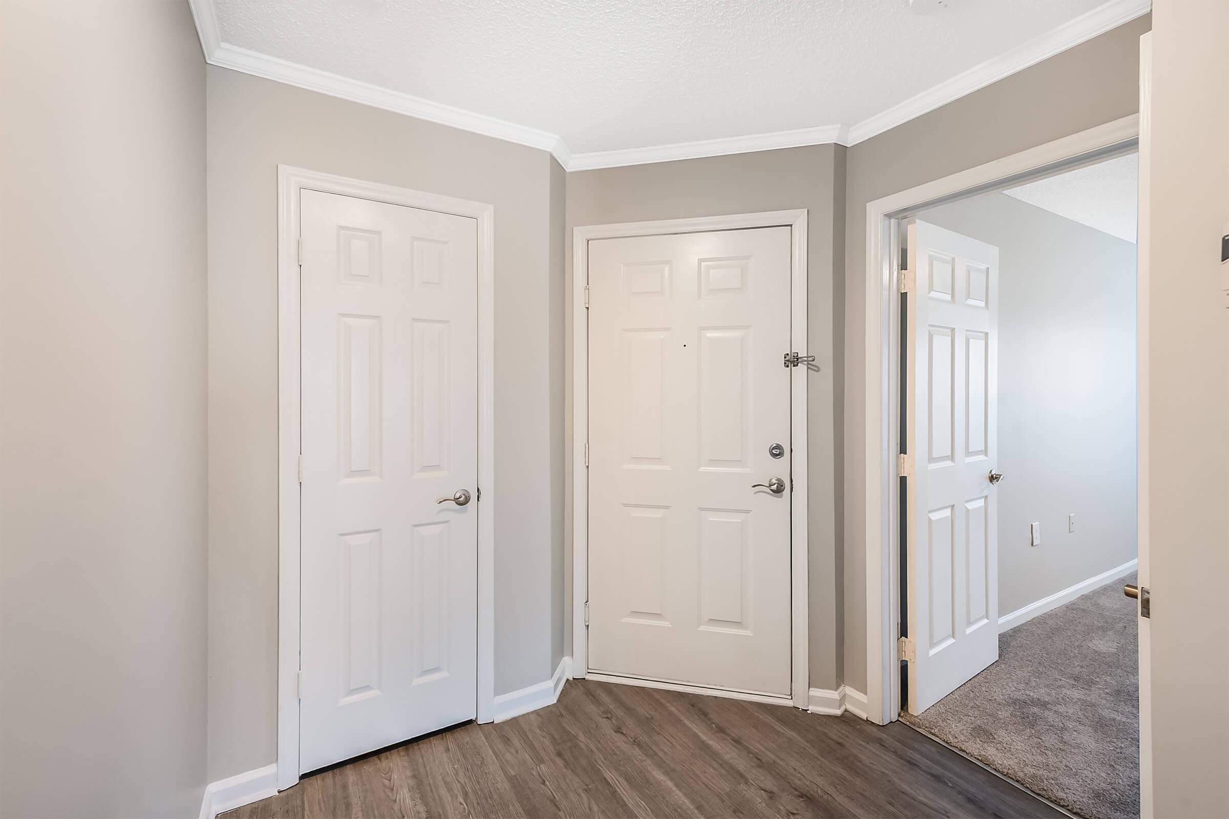 A well-lit entryway featuring two closed doors on the left and a single door on the right. The walls are painted in a neutral color, and the flooring is a light wood. A corner of the room is visible, showcasing the clean, modern design of the space.