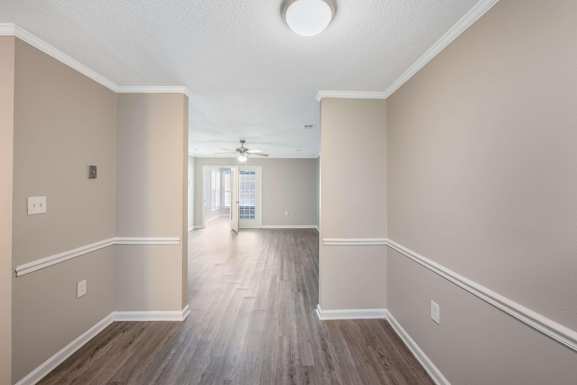 A spacious hallway leads to a brightly lit living area, featuring beige walls and laminate flooring. The ceiling has a light fixture, and there is a ceiling fan visible in the adjacent room. Large windows in the living area allow natural light to flow in.