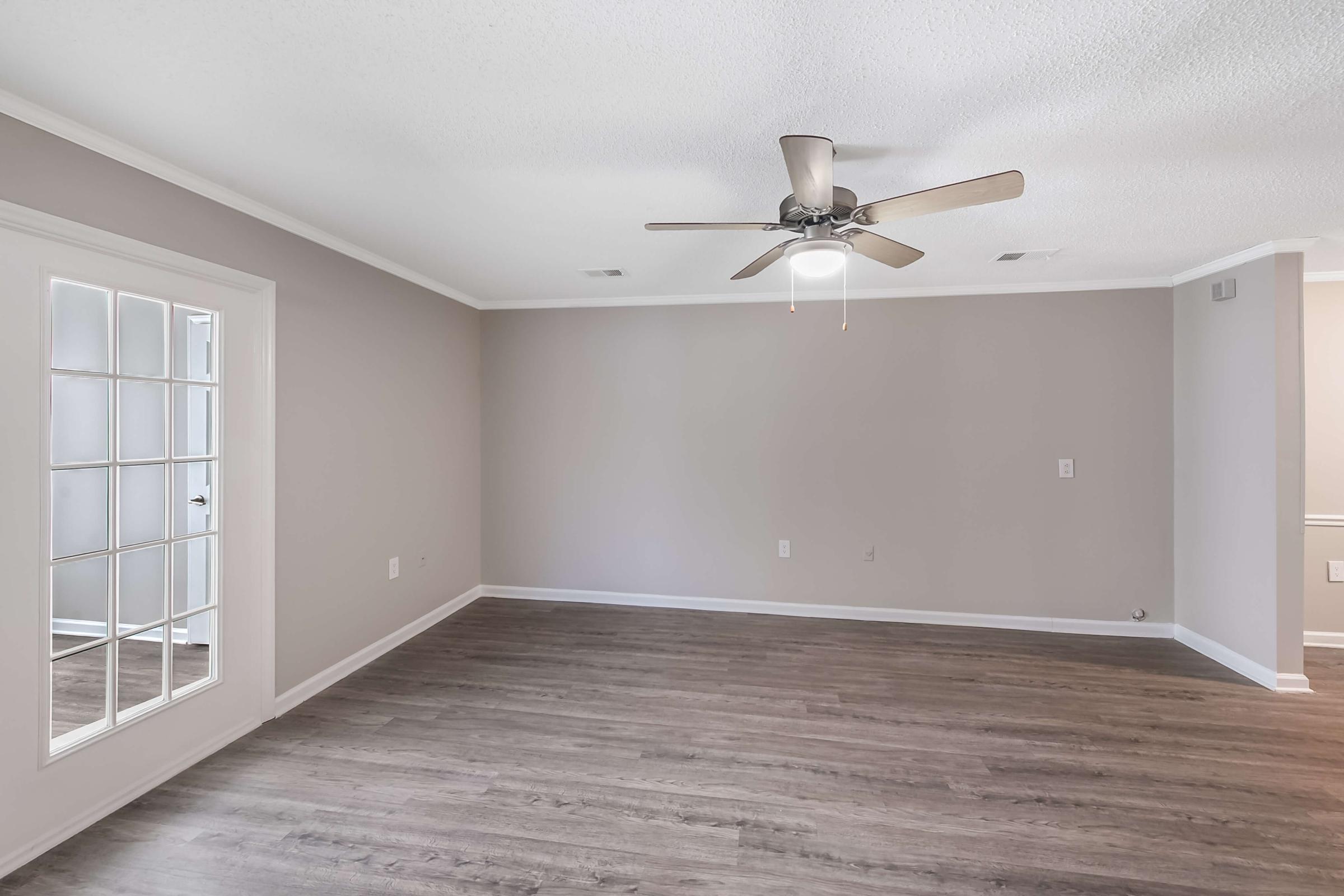 Spacious living room with light gray walls, a ceiling fan, and laminate flooring. A set of French doors on the left leads to another room, while natural light fills the area from a window. The room is empty, ready for furniture arrangement.