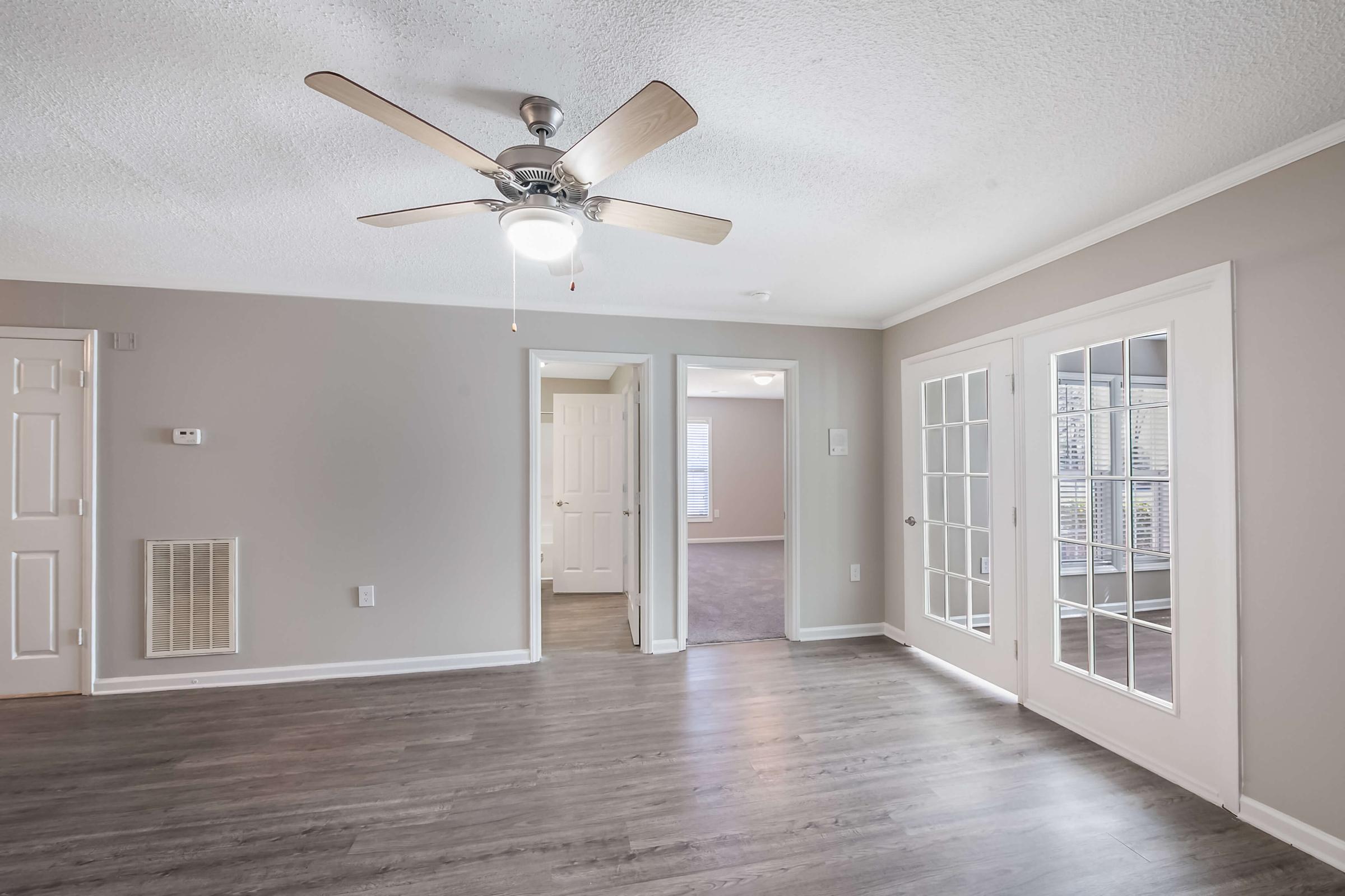 A spacious living area featuring a ceiling fan, light-colored walls, and laminate flooring. The room includes a doorway leading to another room and glass-paneled French doors that provide a view into an adjacent space. Natural light filters through the windows, enhancing the openness of the area.