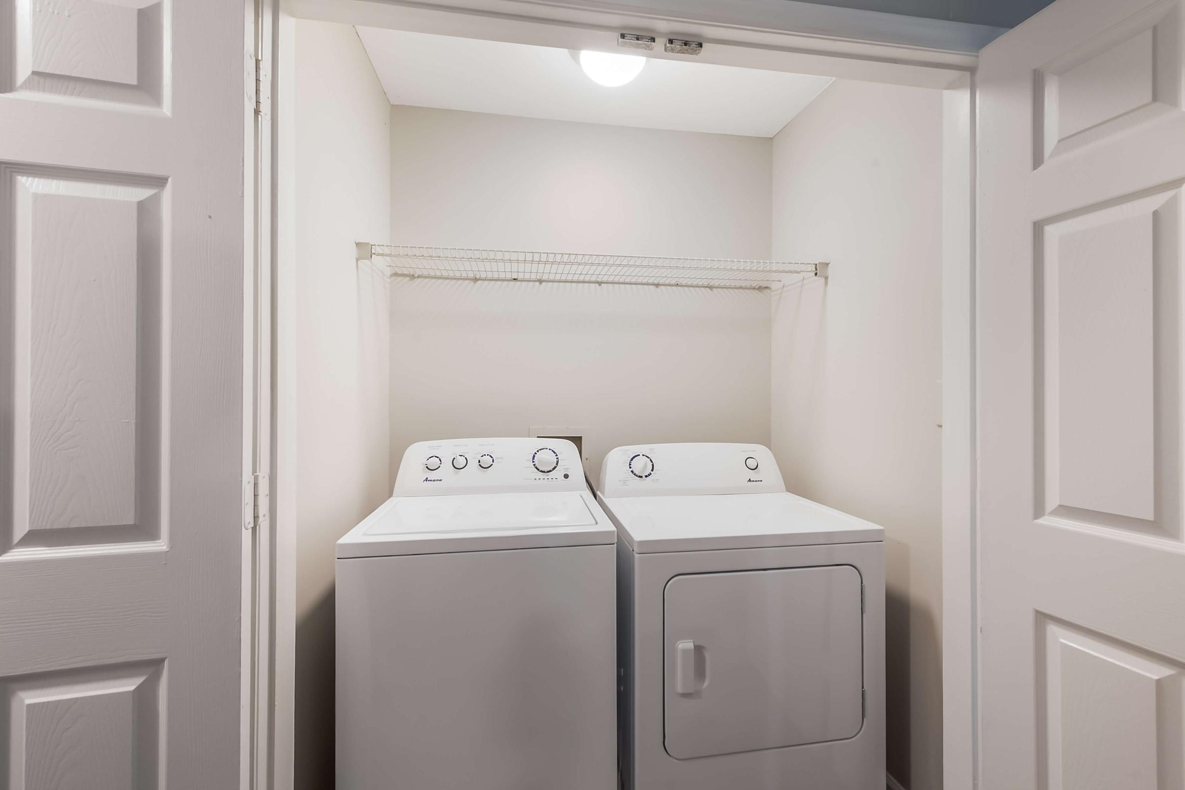 A laundry closet featuring a stacked washer and dryer. The appliances are white and positioned side by side, with a wire shelf above them. The room has beige walls and is illuminated by a ceiling light, with double doors partially open to reveal the space.