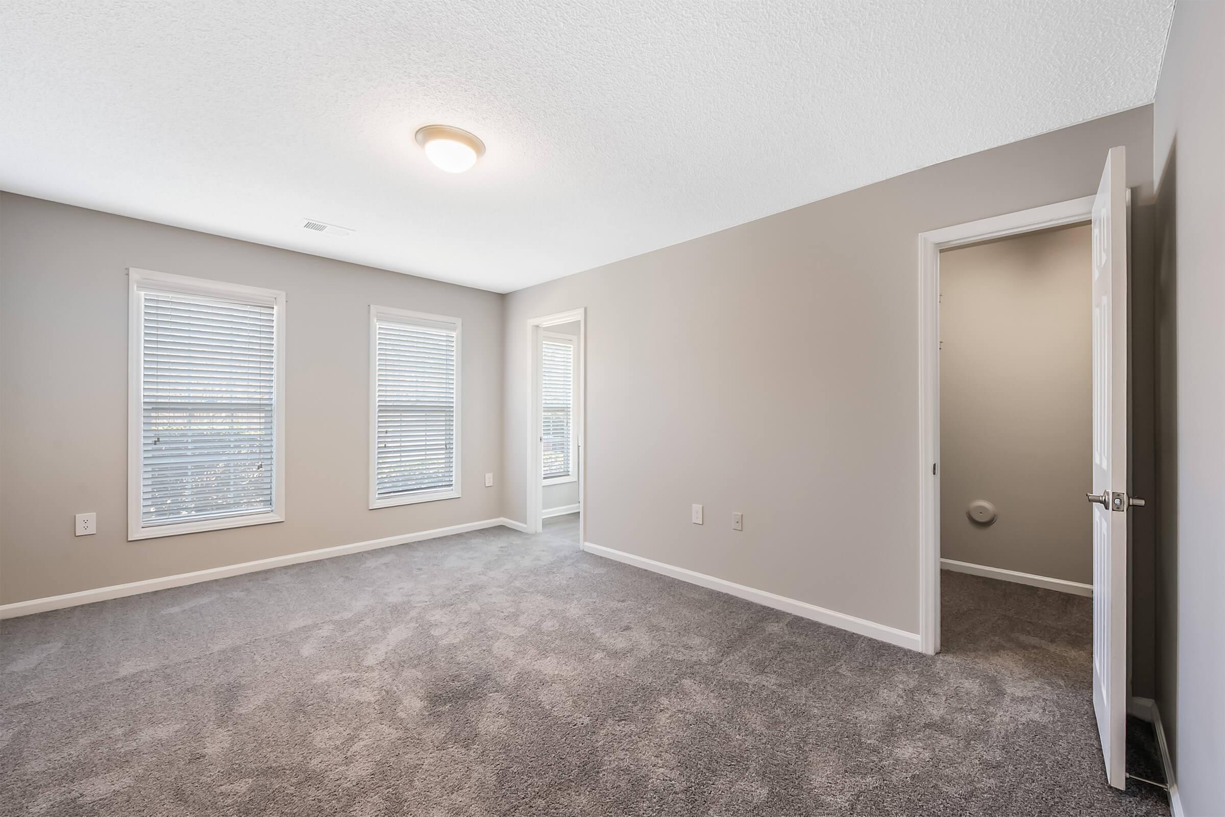 Empty bedroom featuring light gray walls and new carpeting. Two windows with blinds allow natural light, and there are two doorways leading to adjacent rooms. The room has a ceiling light fixture and a clean, spacious feel.