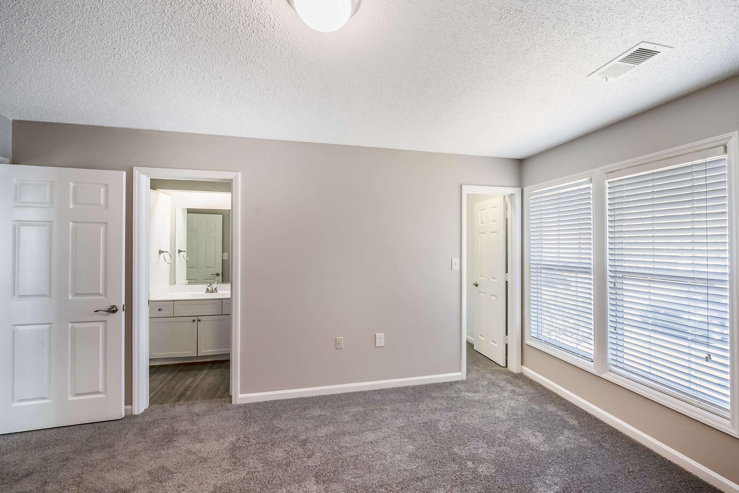 A spacious bedroom with light gray walls and plush carpet. There are two closed doors, one leading to a bathroom with a vanity visible, and the other leading to another room. Large windows with white blinds provide natural light, enhancing the bright and airy atmosphere of the room.