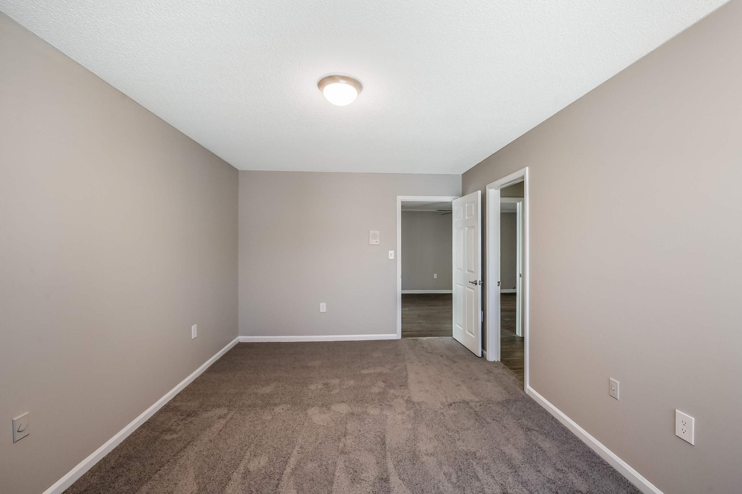 A spacious room with light gray walls and plush, beige carpet. It features a ceiling light and two doorways leading to adjacent areas. The room is empty, creating an airy and open feel, with no furniture present.
