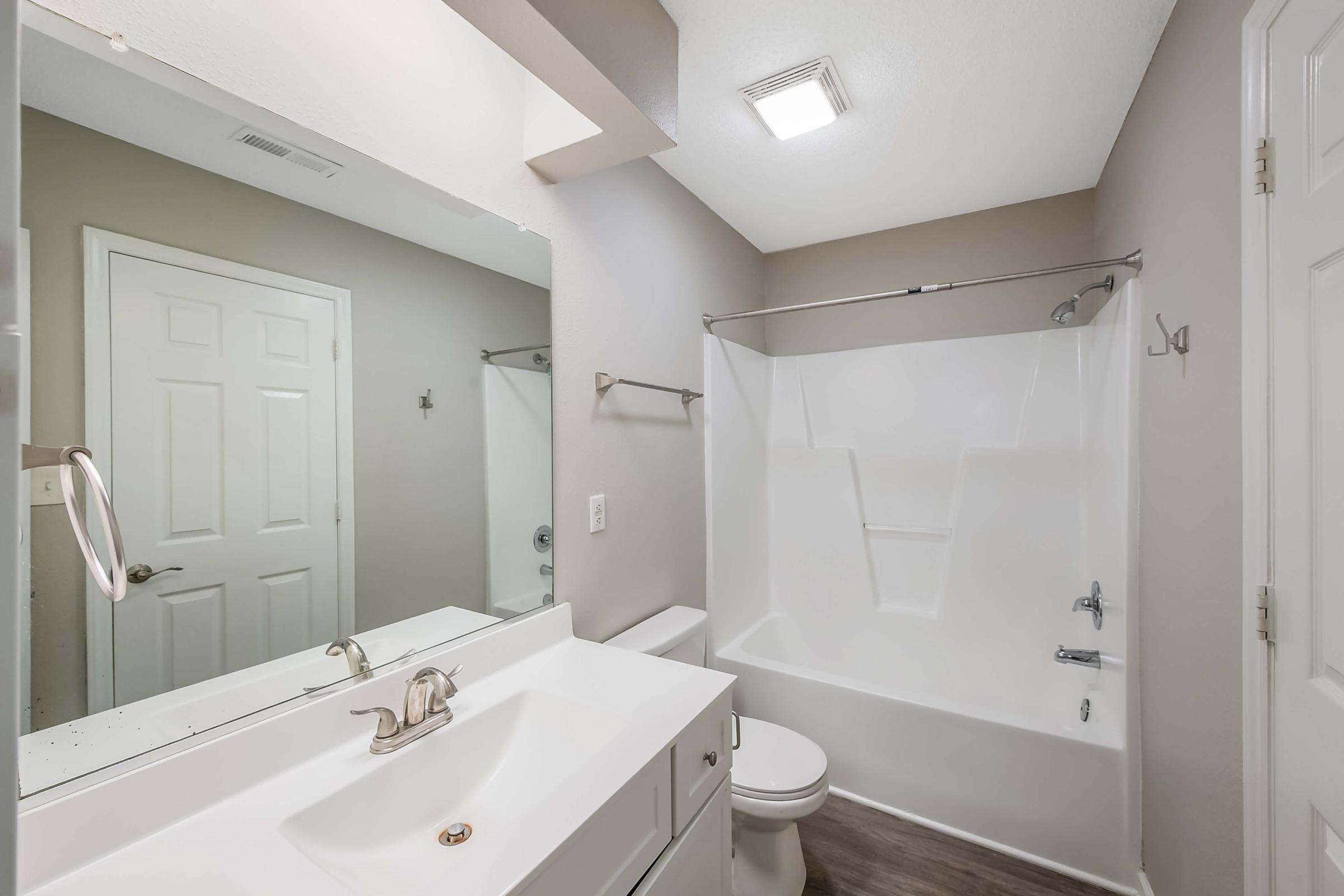A clean bathroom featuring a white bathtub with a shower curtain, a white vanity with a sink, a large mirror, and light-colored walls. The flooring is laminate, and there are white cabinet doors and a towel bar mounted on the wall. The space is well-lit with ceiling lighting.