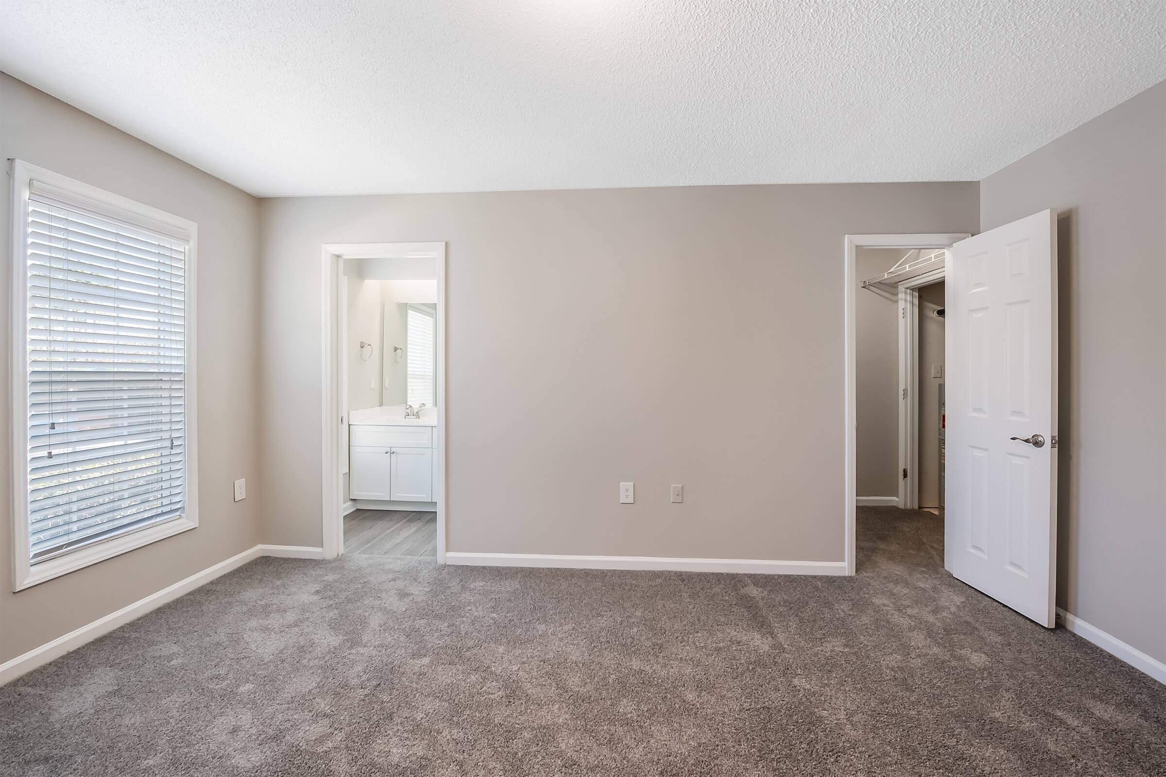 A spacious, empty bedroom featuring light gray walls and carpet. Natural light comes through a window with white blinds. Two doors lead to adjacent rooms, one possibly a bathroom with a white vanity visible, and the other might lead to a closet.