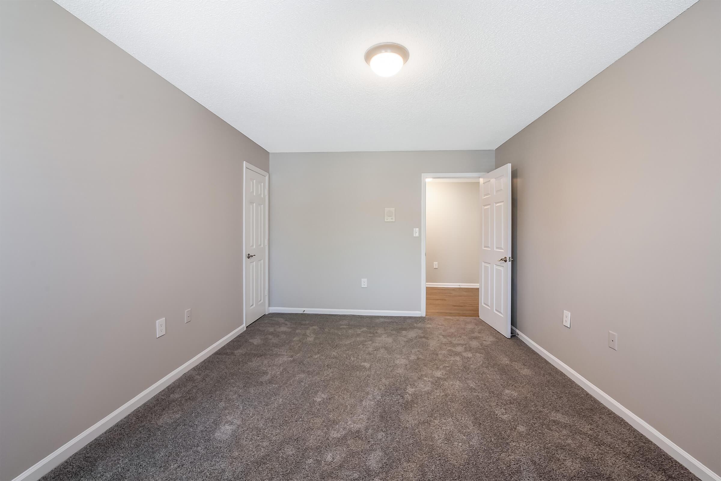 Interior view of a vacant room with light gray walls and plush carpet. The space features a central ceiling light, two closed doors leading to other rooms, and a doorway at the far end. The overall ambiance is neutral and spacious, suitable for various uses.