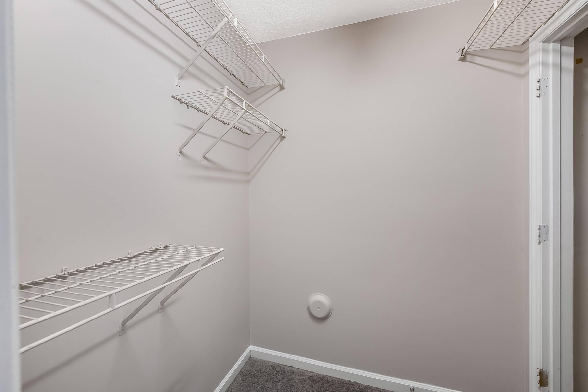 A simple, empty closet with light gray walls and several wire shelving units mounted on the walls. The floor is carpeted, and there is a circular vent on one wall. The overall space appears clean and organized, suitable for storage.