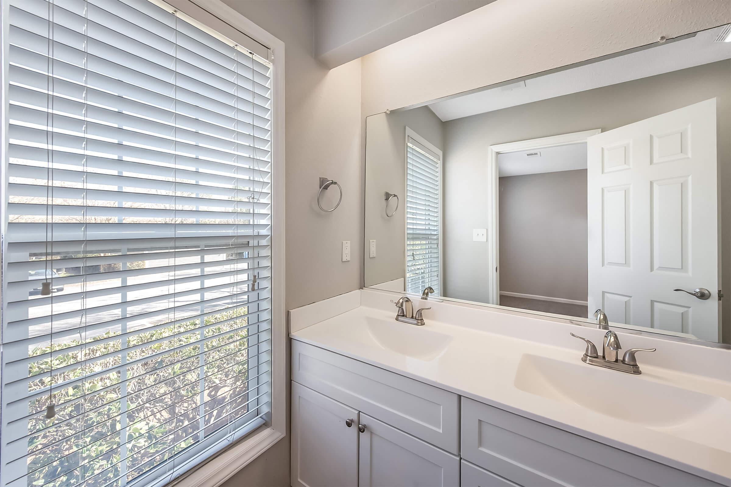A modern bathroom with dual sinks, a spacious mirror above the sink, and a window featuring horizontal blinds. The walls are painted in a light gray color, and there's an open door leading to another room. The overall ambiance is bright and clean.