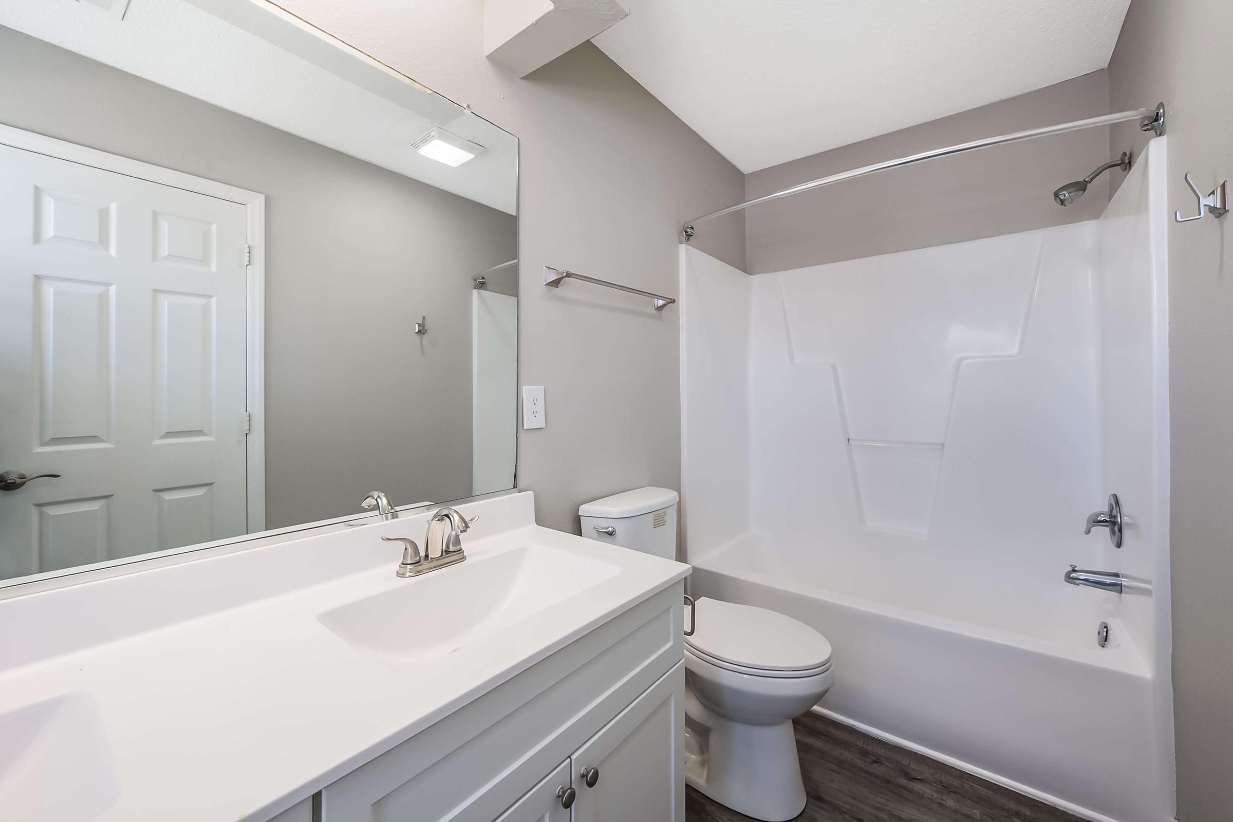 A modern bathroom featuring a white bathtub and shower combination, a toilet, and a double sink vanity with a large mirror. The walls are painted in a light gray, and the floor is a dark laminate. There is a closet door visible in the background. The overall design is clean and minimalist.