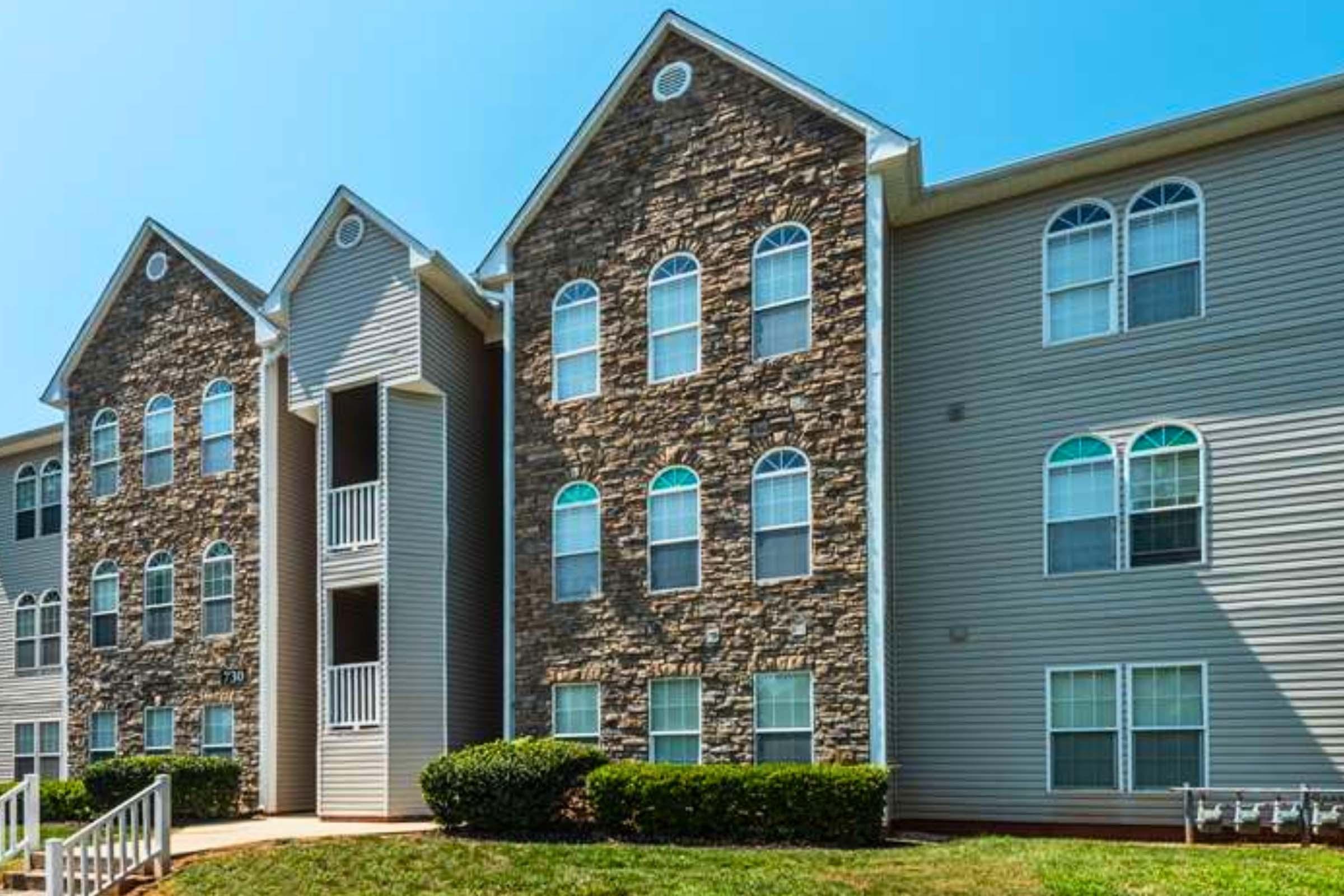A multi-story apartment building featuring a combination of stone and vinyl siding. The structure has several large windows and a symmetrical design, with green grass and shrubs in the foreground, under a clear blue sky.