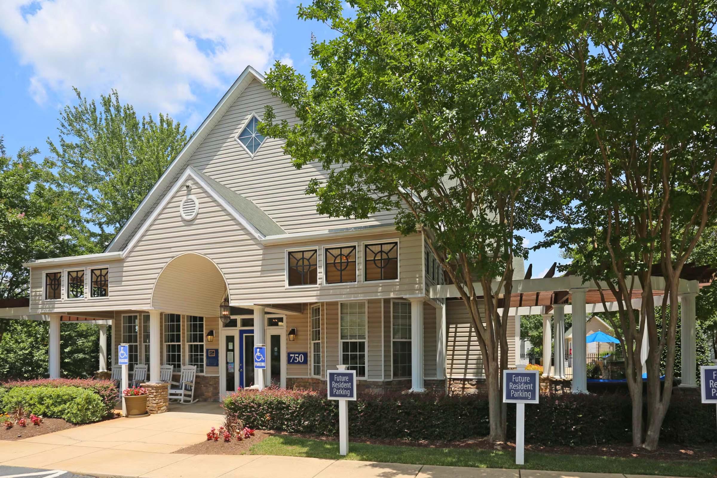 A light-colored, one-story building with a peaked roof and decorative windows. It features an accessible entrance, surrounded by greenery and a well-maintained landscape. Signs are placed outside indicating parking. The scene is bright and sunny, showcasing a welcoming atmosphere.