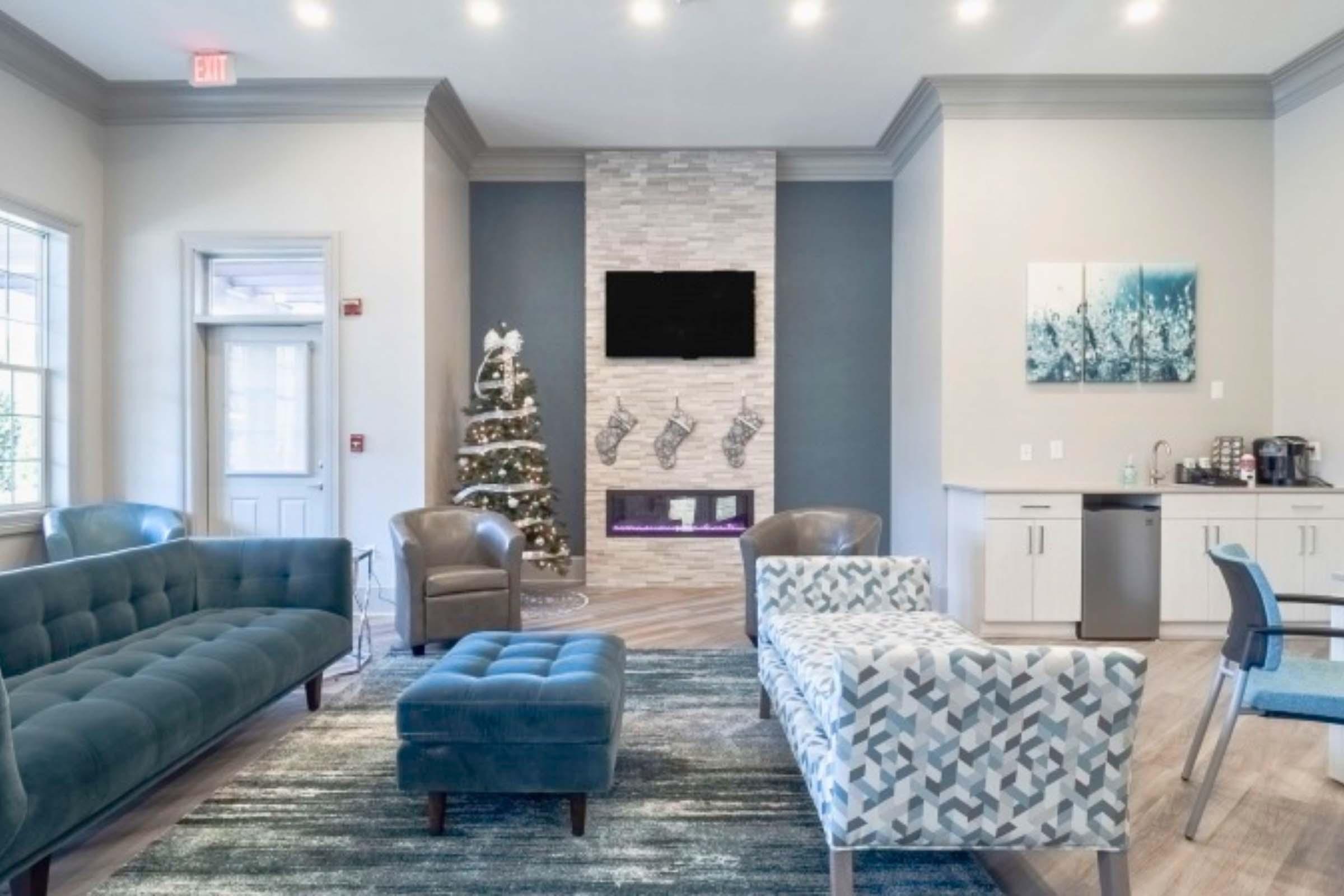 A cozy living area featuring a blue sectional sofa, a patterned chair, and an ottoman. The decor includes a Christmas tree and stockings above a modern stone fireplace. A kitchenette is visible in the corner, and soft lighting enhances the inviting atmosphere.