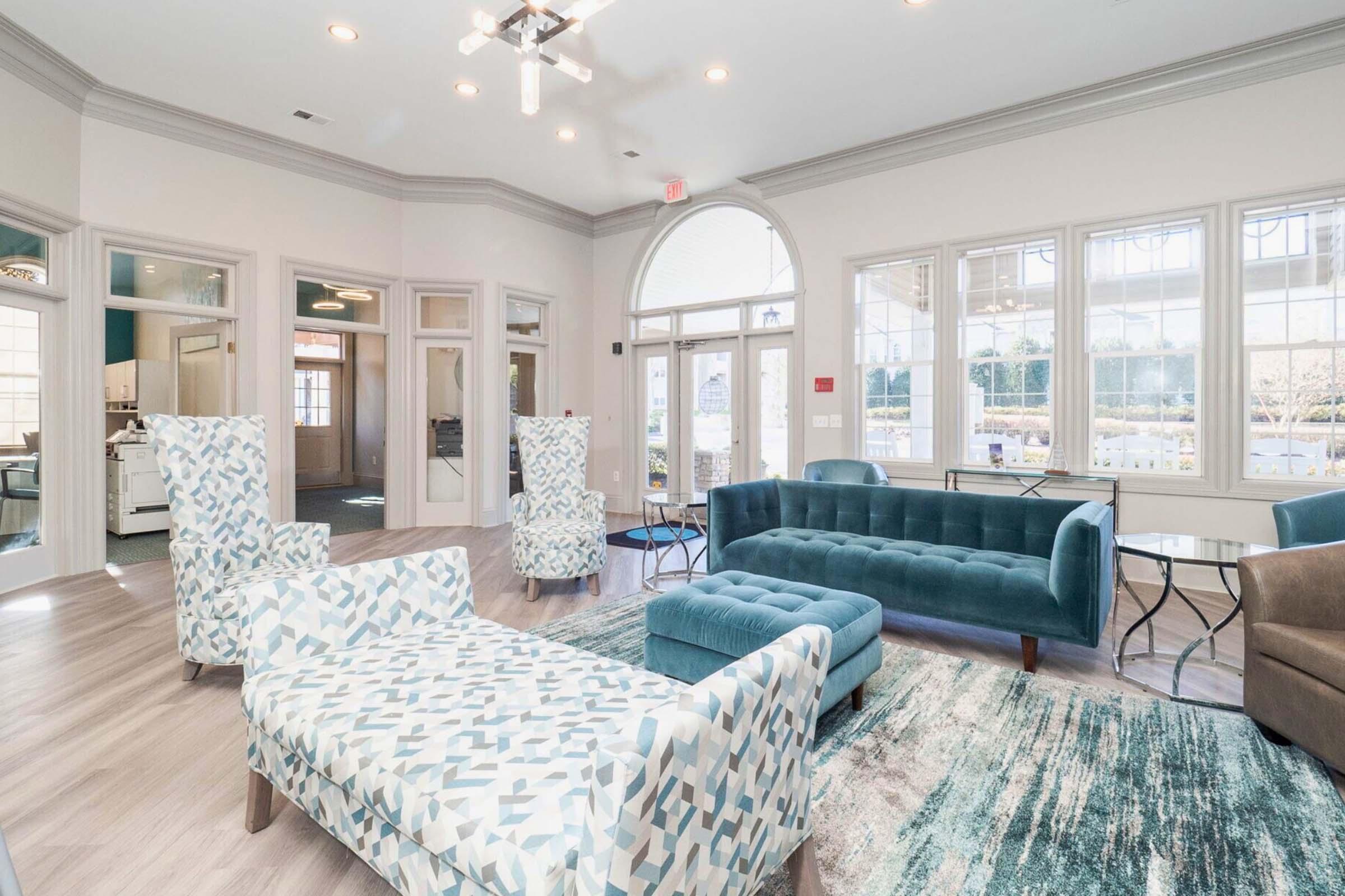 A bright, modern lounge area featuring two patterned armchairs, a teal tufted sofa, and a light-colored rug. Large windows allow natural light to fill the space, showcasing a welcoming atmosphere with neutral walls and stylish decor.