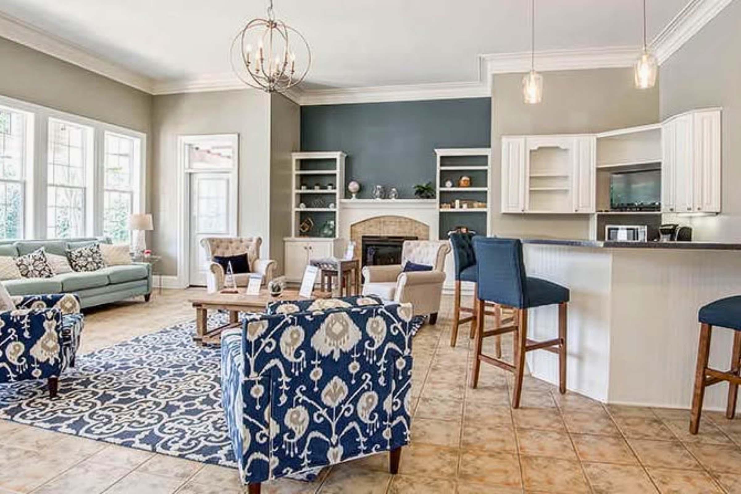 Bright and inviting living space featuring a light-colored sofa, two patterned armchairs, and a coffee table. The room includes a fireplace, built-in shelves, and large windows that let in natural light. A kitchen bar area with high stools is visible, along with decorative lighting fixtures hanging from the ceiling.